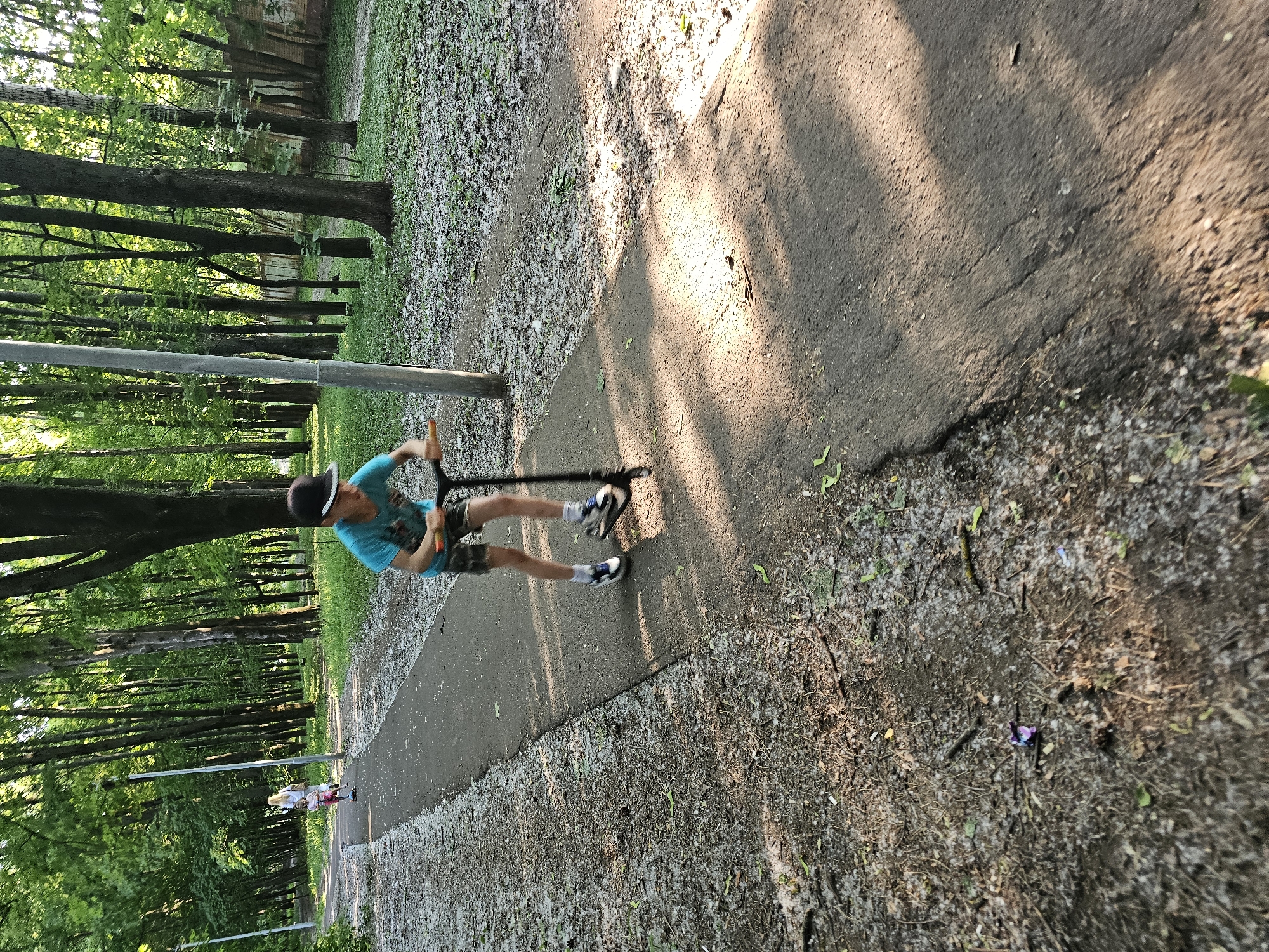 In Khimki, Russia, a young boy with dark hair, wearing a blue t-shirt, camouflage shorts, white socks with blue stripes, and black and white sneakers, rides a black scooter down a paved path in a park. He is wearing a black baseball cap. In the background, further down the path, a woman in a white outfit and a young girl on a pink scooter are visible. The park is characterized by a dense line of tall trees with green foliage, and the ground is covered with a mixture of dirt, leaves, and white debris, possibly poplar fluff. Sunlight casts dappled shadows on the path. A lamppost is situated to the left of the path.