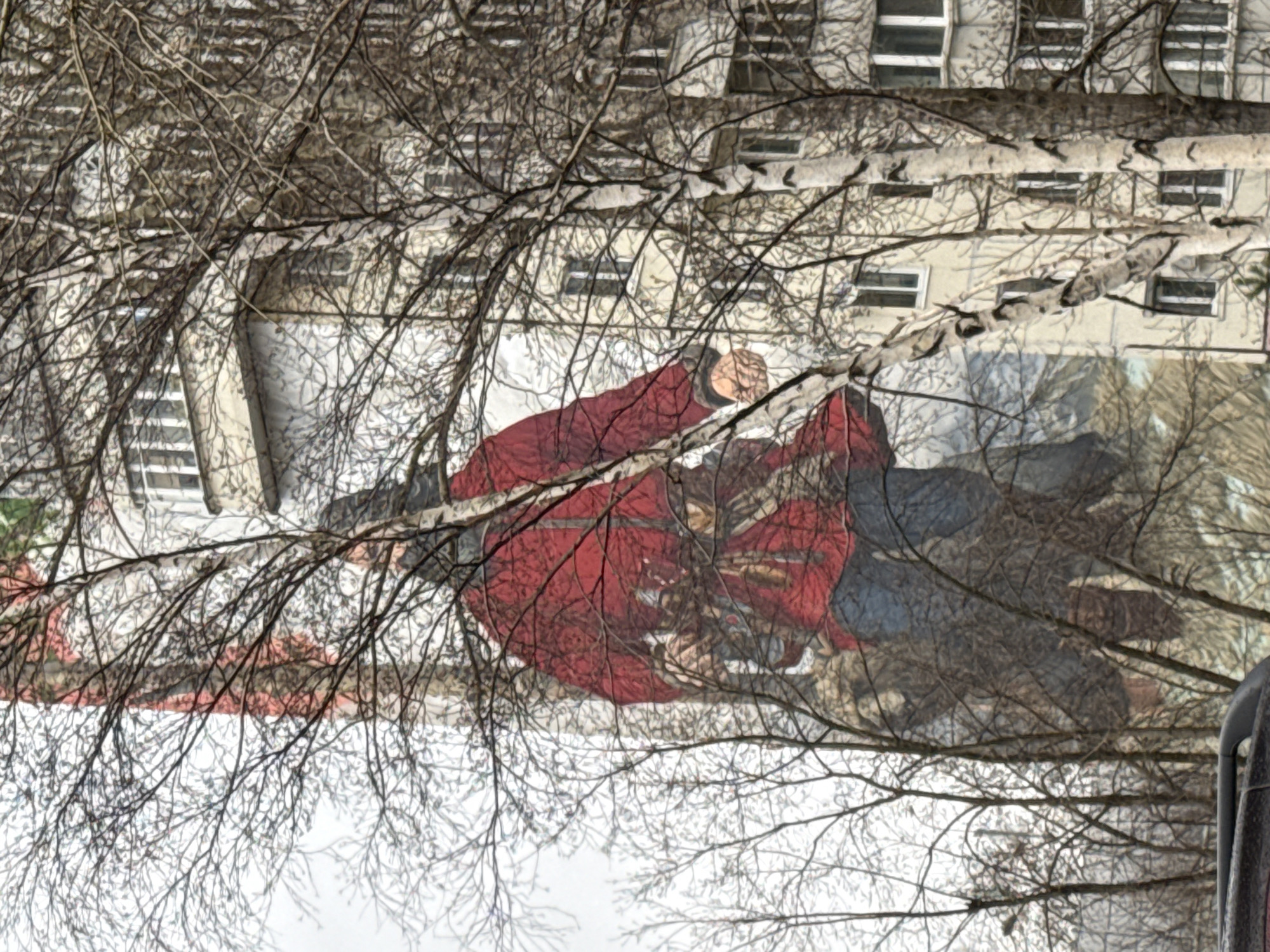 The image is a shot of a group of people in front of a building in Perm, Russia. The building is a light-colored, multi-story structure with rows of windows. Bare tree branches partially obscure the building and people. In the foreground, three or four people are seen, one of whom is wearing a bright red coat. The people are interacting with one another. The overall tone of the image suggests it was taken in a cold or winter setting, given the bare trees. The scene is urban, captured during the daytime.
