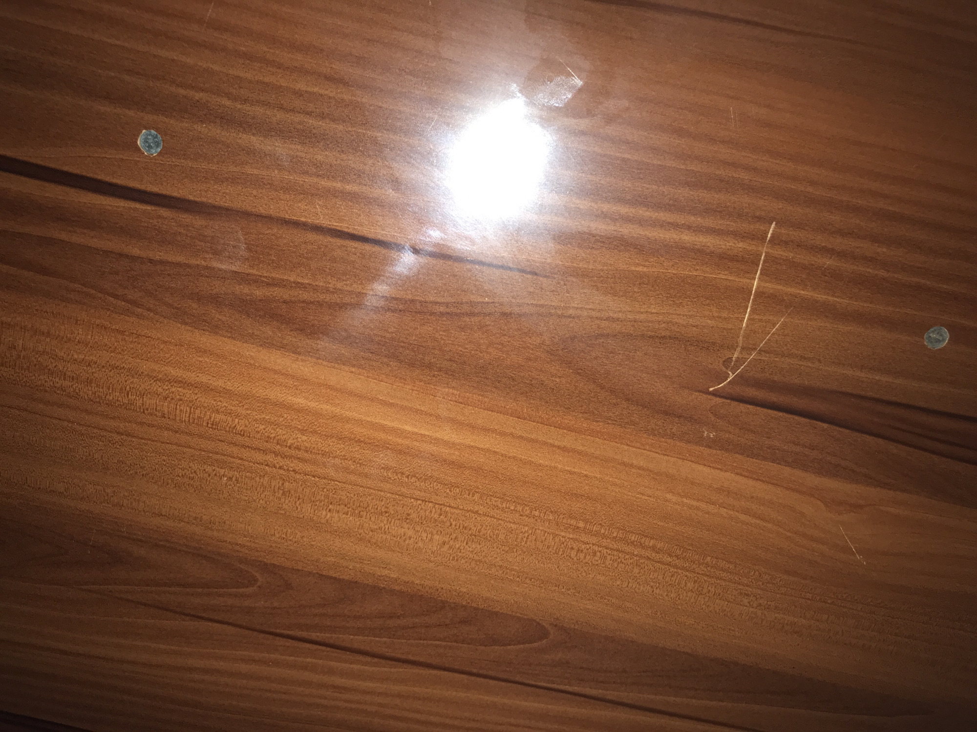 The image shows a close-up, high-angle view of a polished wooden surface, likely a table or furniture. The wood has a warm, reddish-brown tone with prominent grain patterns running vertically. There are several imperfections visible on the surface. Two small, circular indentations are present, one near the top right and another near the bottom center. A bright, reflective glare from a light source dominates the mid-right portion of the image. A fine, light-colored hair or thin strand of material is visible extending across the wood grain in the lower middle section. Several faint, thin scratches are also present, particularly in the lower half of the image. The overall impression is that of a well-used wooden surface. There are no people or clear indicators of a specific location within Jos, Nigeria.