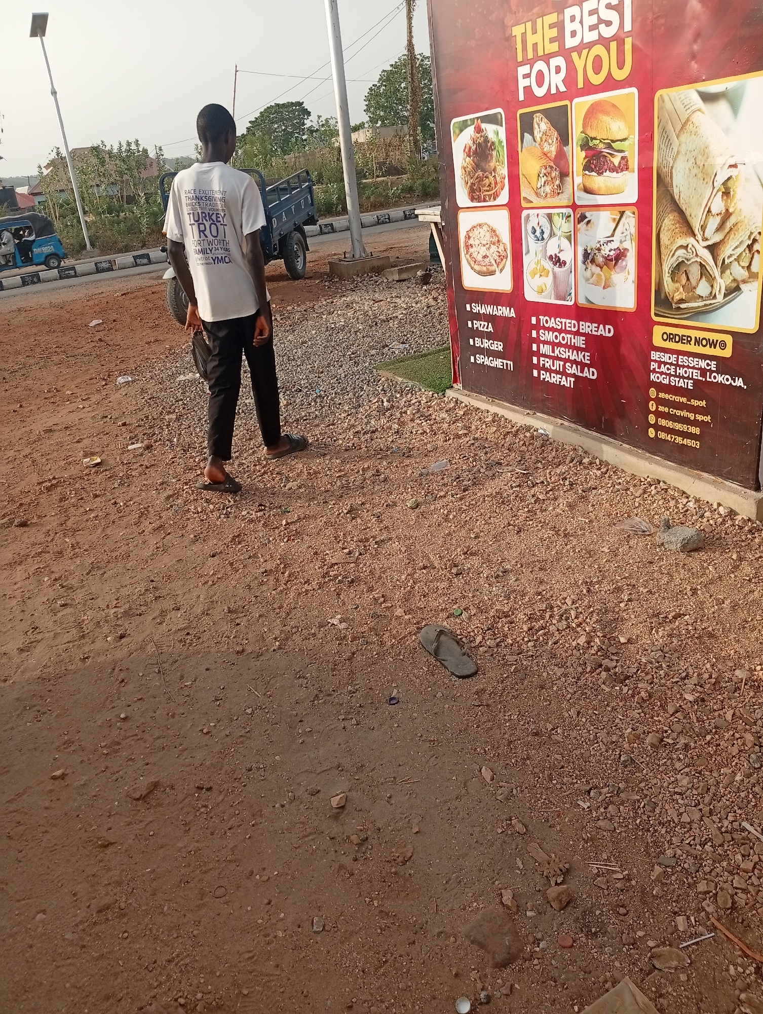 A medium shot captures a young man, seen from the back, standing on a gravelly, reddish-brown patch of ground. He is dressed in a white T-shirt with text and black pants, with sandals on his feet. To his right stands a large advertisement board advertising a food business, with items such as shawarma, pizza, burger, spaghetti, toasted bread, smoothie, milkshake, fruit salad, and parfait listed. The board also displays an "Order Now" message, followed by the location "Beside Essence Place Hotel, Lokoja, Kogi State," indicating the business's location in Adankolo, Nigeria. Contact details including social media handles and phone numbers are printed. Behind the man and the advertisement, a three-wheeled motor vehicle is visible on the road. The environment includes utility poles and some distant vegetation, under a bright, overcast sky. A single sandal rests on the ground at the bottom of the frame.