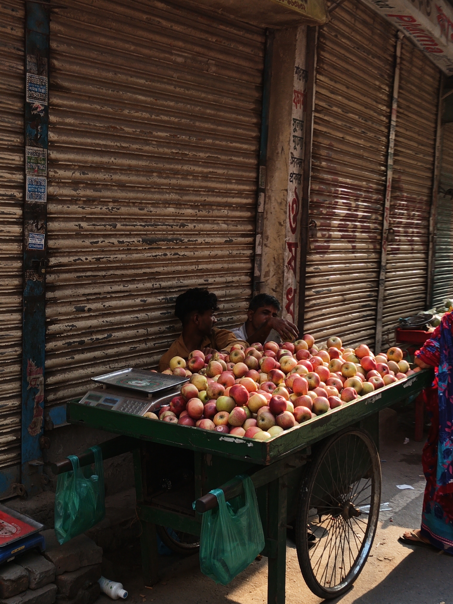 Two boys sit behind a cart laden with red and yellow apples, likely in Dhaka, Bangladesh. The cart is green and has a wheel on one side. A woman stands to the right, partially visible, wearing a colorful garment. The backdrop consists of closed, textured metal shopfronts. A weighing scale sits on top of the apples. Green plastic bags hang from the sides of the cart.
