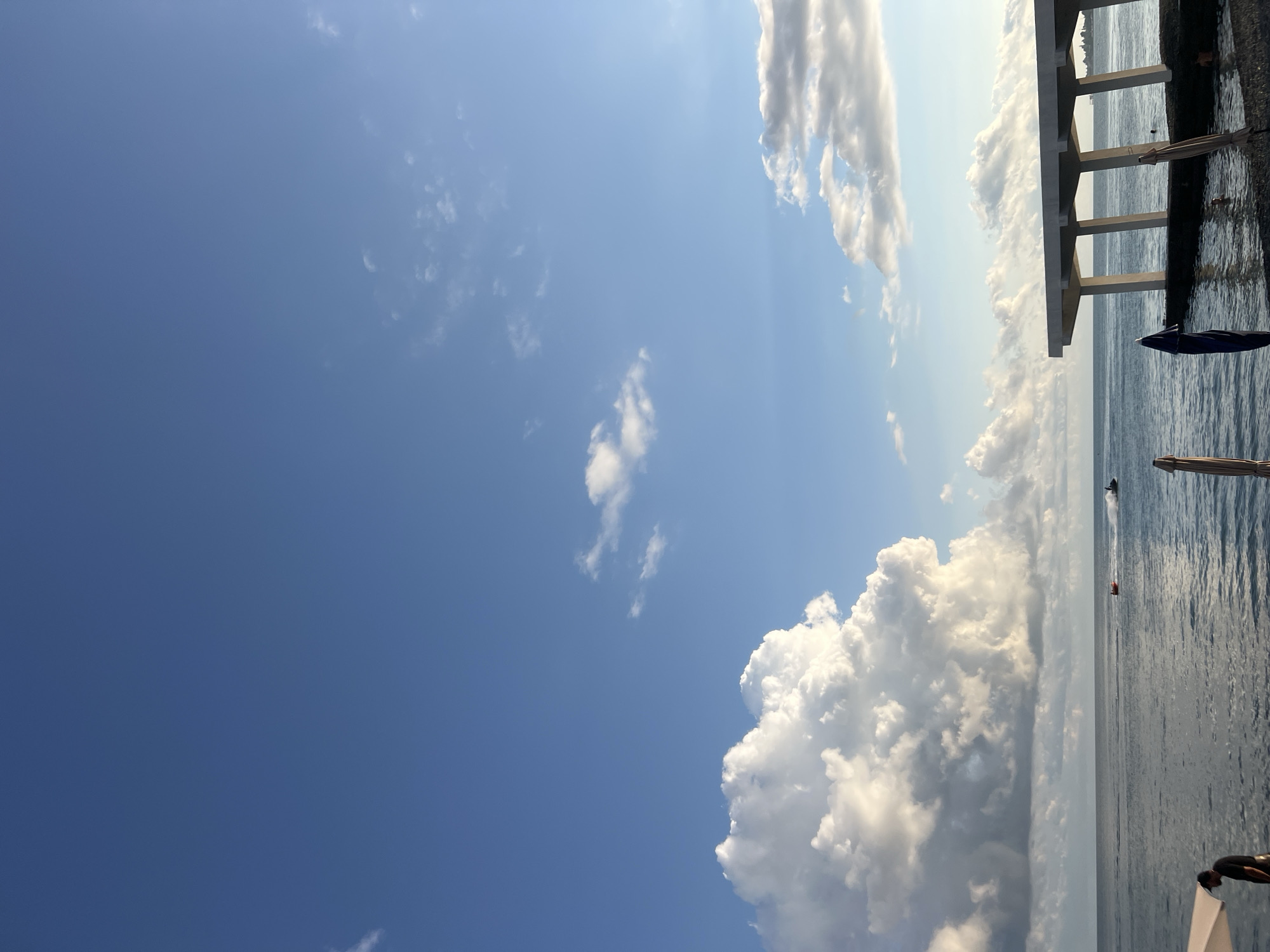A wide-angle, low-angle shot captures a serene seascape with a clear blue sky dominating the upper portion. The horizon is marked by the calm, dark blue ocean, meeting a distant, hazy shoreline. On the left side of the frame, a massive, fluffy white cloud formation stands out against the blue expanse, its illuminated edges suggesting late afternoon sunlight. Smaller, scattered clouds drift across the sky.
In the middle ground, the ocean is dotted with activity. A bright orange inflatable tube being pulled by a jet ski moves from left to right, leaving a white wake. Further to the right, a black jet ski is also in motion, creating a similar trail on the water.
The foreground features a man standing near the water's edge on the left, wearing a dark t-shirt and a baseball cap, partially obscured by a white umbrella structure. Several closed beach umbrellas, some brown and one dark blue, are positioned along the shore. To the right, a modern, minimalist white structure with a flat roof supported by angular columns extends over the water. It appears to be a pavilion or a viewing platform. The water around the pavilion is clear enough to see people swimming. The shoreline is a dark-colored pebble beach. The overall impression is one of a tranquil day at the beach, likely in a resort area. The scene is likely set in Sokhumi, Abkhazia, given the context.