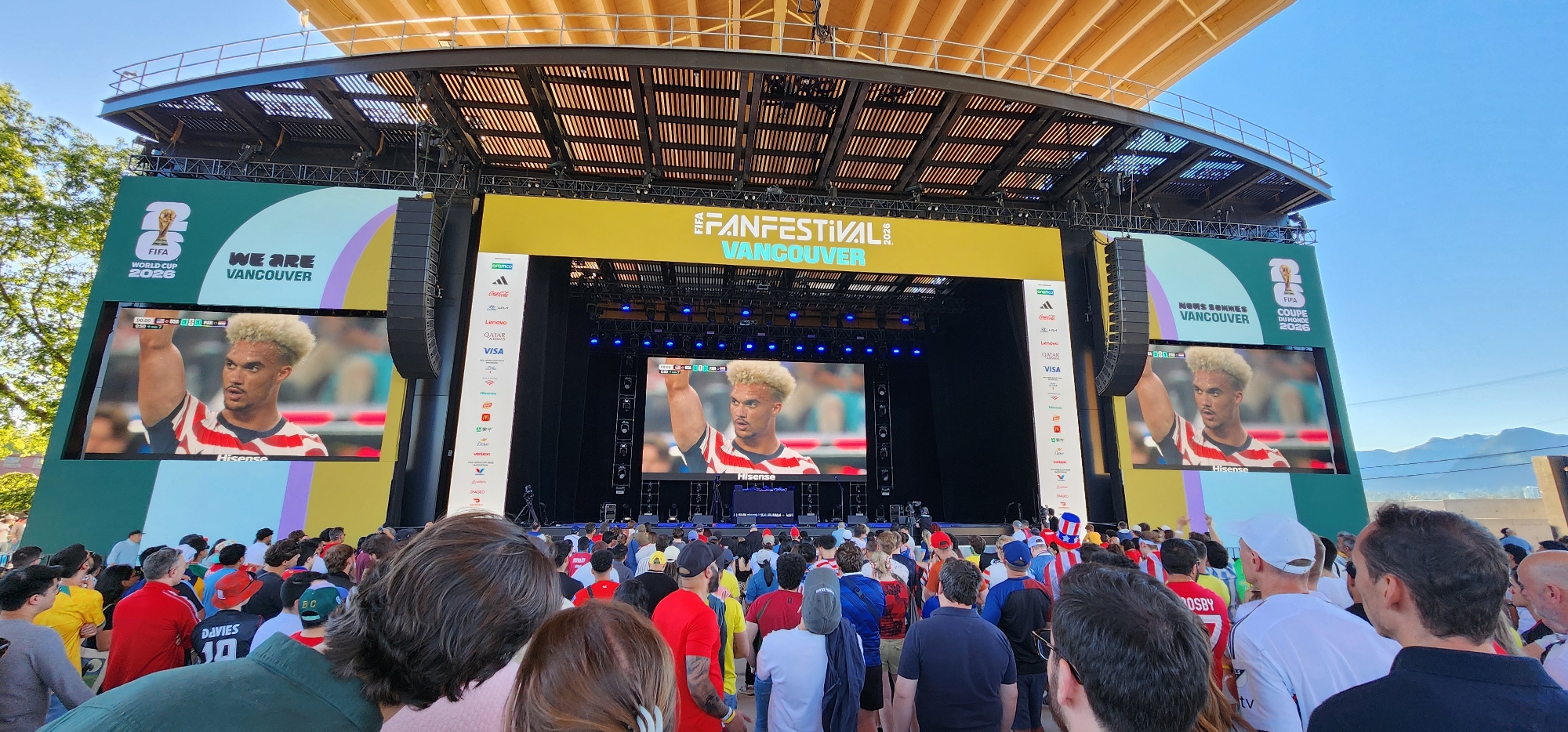 The image shows a large outdoor screen displaying a soccer player with blond hair, wearing a red, white, and blue jersey with the name "Hisense" visible on the screen. Above the soccer player's image, the text "FIFA FANFESTIVAL VANCOUVER 2026" is displayed in yellow. On either side of the central screen, two smaller screens show the same soccer player. The left screen has text "FIFA WORLD CUP 2026 WE ARE VANCOUVER" written in green. The right screen has text "NOUS SOMMES VANCOUVER FIFA COUPE DU MONDE 2026". A crowd of people is gathered in front of the screens, facing the display and appearing to be watching the event. The scene is set against a backdrop of blue sky and distant mountains. The overall environment suggests a public viewing event for a soccer tournament.
