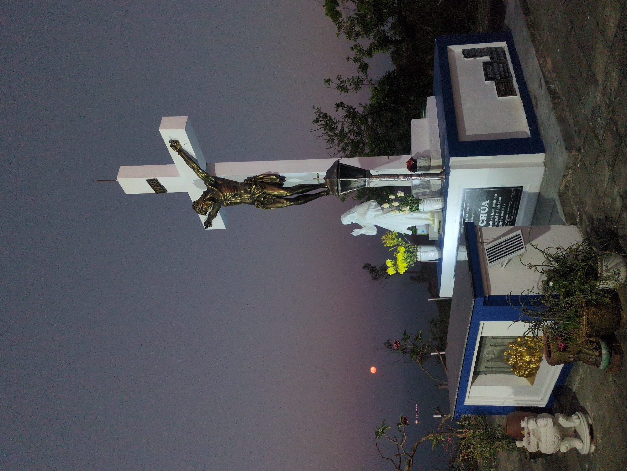 The image captures a grave in what appears to be a cemetery. The scene is dominated by a white cross adorned with a golden figure of Jesus Christ. A statue of the Virgin Mary and vases of flowers are placed at the base of the cross. The grave itself is constructed with a blue and white structure, featuring a plaque with inscriptions in Vietnamese, including the word "CHÚA". A golden statue is visible within a lower section of the grave structure. Potted plants and a small white statue of a lion decorate the surrounding area. In the background, the sky transitions from a darker tone to a lighter, reddish hue near the horizon. Based on the OCR, the location is in the city of South Quy Nhon Ward, Vietnam.