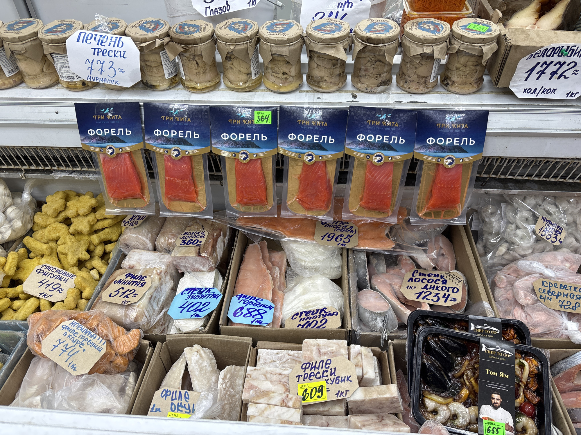 The image shows a refrigerated display case at a fish market in Izhevsk, Russia. The case is filled with various fish and seafood products, each with price tags written in Russian. At the top, there are jars of cod liver pate, next to packages of trout fillets. Below these, are various types of fish fillets and steaks, including cod, pollock, pink salmon and chum salmon. The bottom shelf contains trays of cooked Tom Yum soup. The prices of each item are prominently displayed. The overall environment suggests a bustling local market.