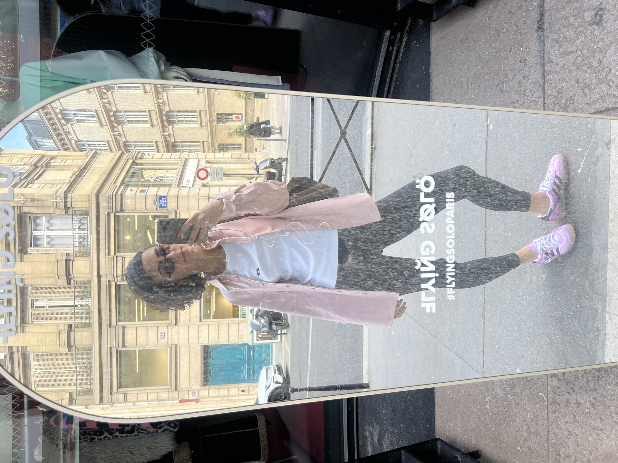 A reflection in a mirror placed outside in Paris, France, depicts a person taking a selfie. The person wears sunglasses, a white shirt, a pink jacket, patterned leggings, and lavender sneakers. The backdrop, visible through the reflection, includes typical Parisian architecture with buildings with multiple floors and ornate details. A street sign is also visible. The mirror has writing on it, presumably advertising information. The setting seems to be a sidewalk next to a storefront, evidenced by glimpses of merchandise inside.