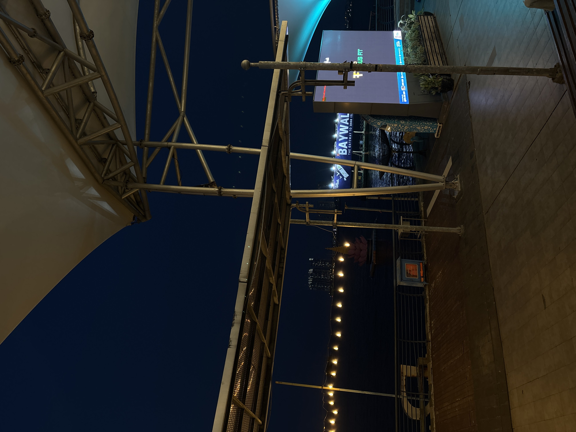 The photograph, taken in North Jakarta, Indonesia, showcases an outdoor commercial area at night. The main subject is the "BAYWAL" sign illuminated in blue and white, advertising "THE GATEWAY TO 1000 ISL." A metal structural frame is visible on the left, supporting a fabric roof element. A string of decorative lights stretches vertically in the distance. The ground is paved and bordered by a brick wall. No people are visible.