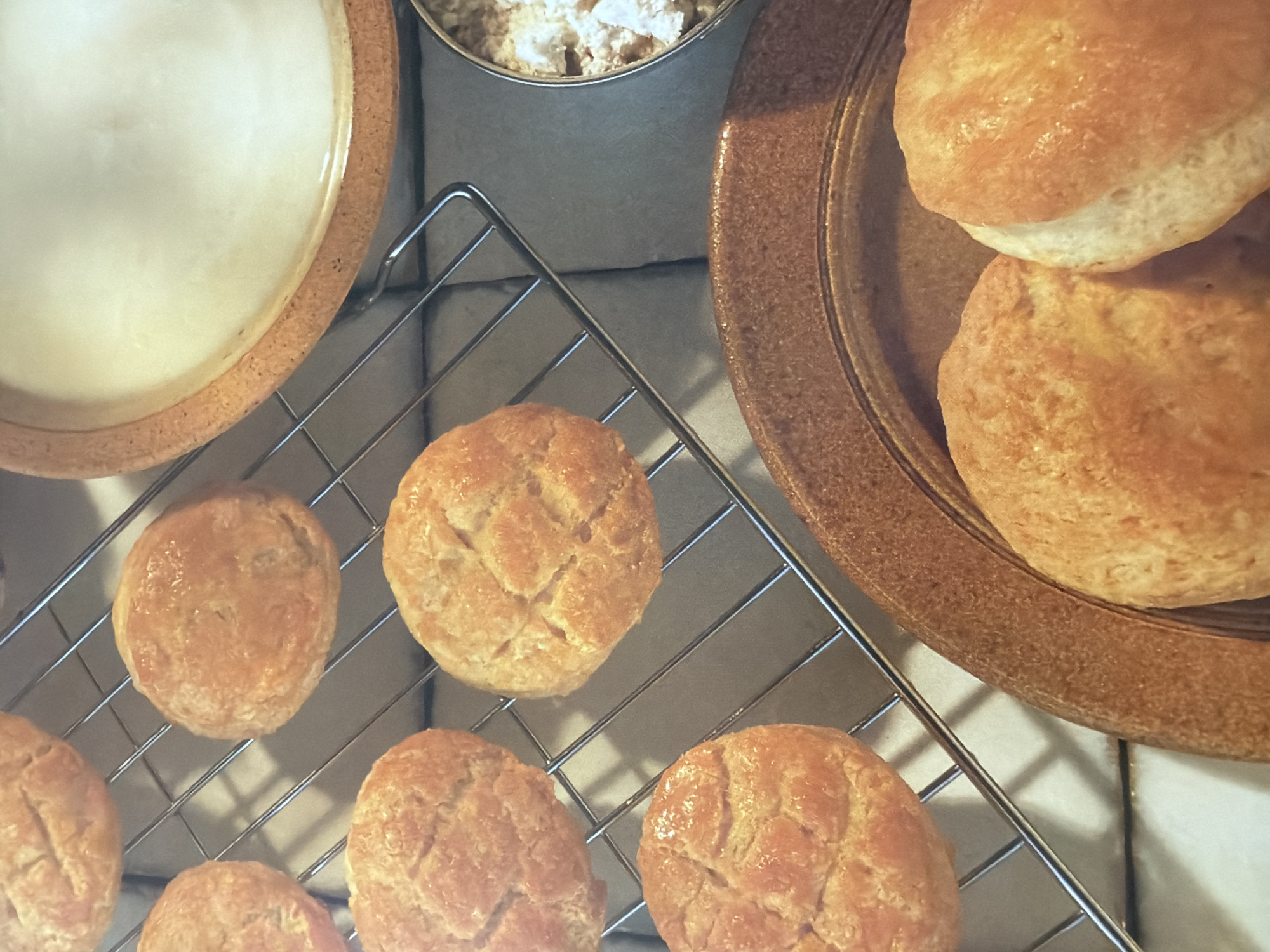 The image shows a close-up, overhead view of freshly baked biscuits on a cooling rack and on a plate. The biscuits are golden brown and appear to be soft and fluffy. Some have a scored pattern on top. To the right of the cooling rack, there is a bowl containing what appears to be shredded cheese or a similar crumbly ingredient, and a larger, shallow bowl filled with a white liquid, possibly milk or cream. In the foreground, two more biscuits are arranged on a rustic, brown plate. The background is a neutral gray surface. The scene is likely set in a kitchen or dining area.