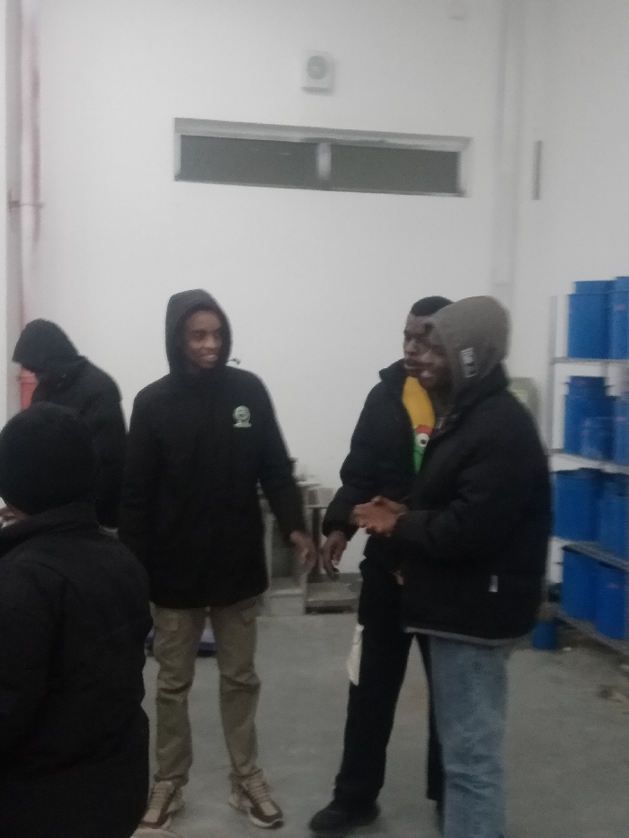 Five people wearing dark hoodies and jackets stand in a brightly lit room. The person in the center wears khaki pants, while another wears blue jeans. Stacks of blue containers are visible in the background to the right. A rectangular window sits high on the back wall, above a light fixture. The overall setting appears to be an industrial or storage space.