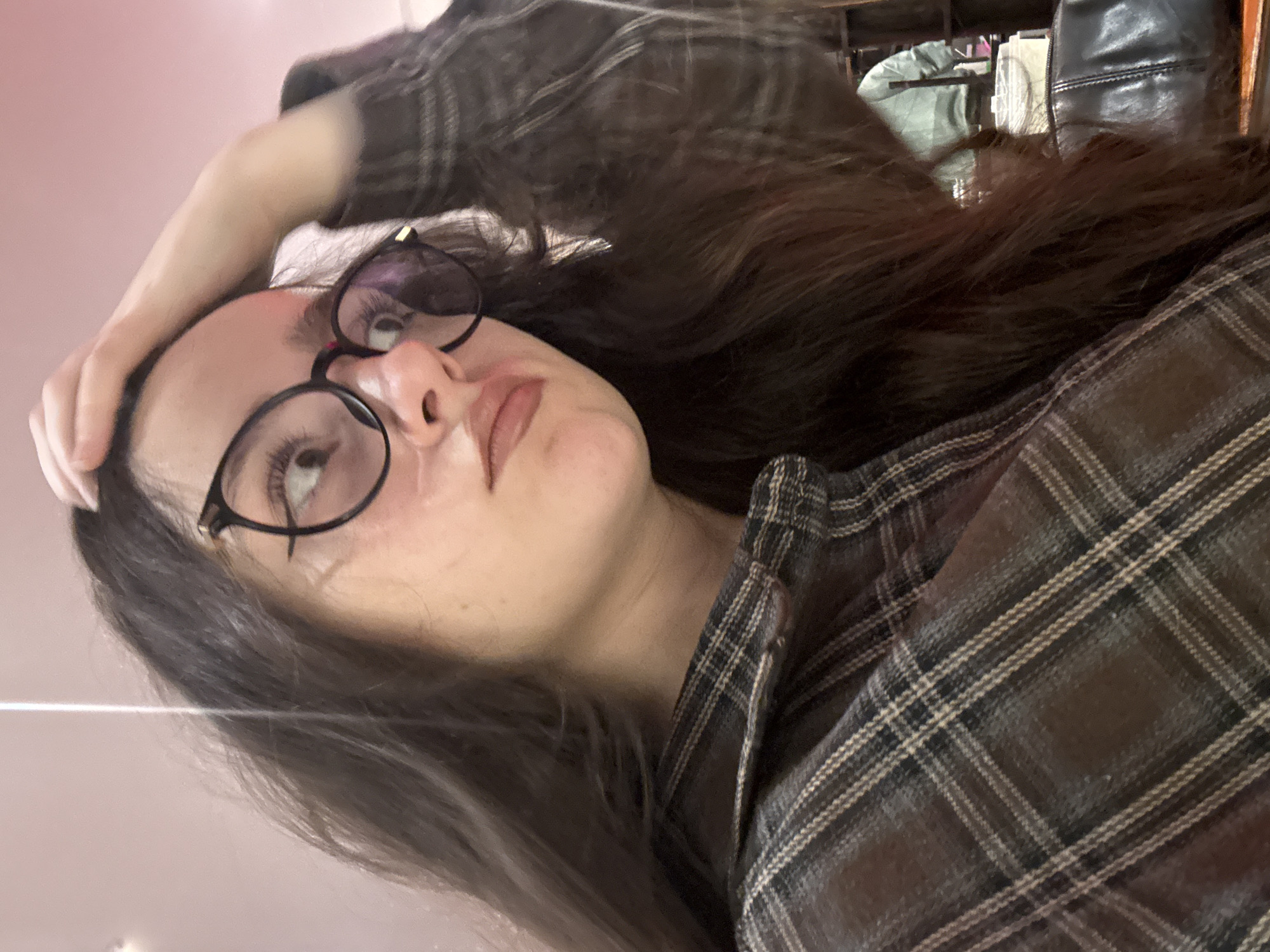 A young female subject is depicted in a close-up, angled shot, occupying the majority of the frame from the chest up. She has long, dark brown hair. She wears round, black-framed eyeglasses with a gold accent visible on the right temple. Her gaze is directed upwards and to the viewer's right. Her lips are slightly pursed, and her eyes are wide, conveying a pensive or questioning expression. Her right hand is raised, with fingers resting on the top of her head. She is dressed in a brown and beige plaid button-down shirt. The background is out of focus, featuring a light pinkish-peach wall or surface on the left. On the right, a blurry figure in a light green garment and a dark, potentially leather, object resembling a bag are discernible. The environment is an indoor commercial or public establishment. No specific visual identifiers are present within the image to ascertain a precise location within Yerevan, Armenia.