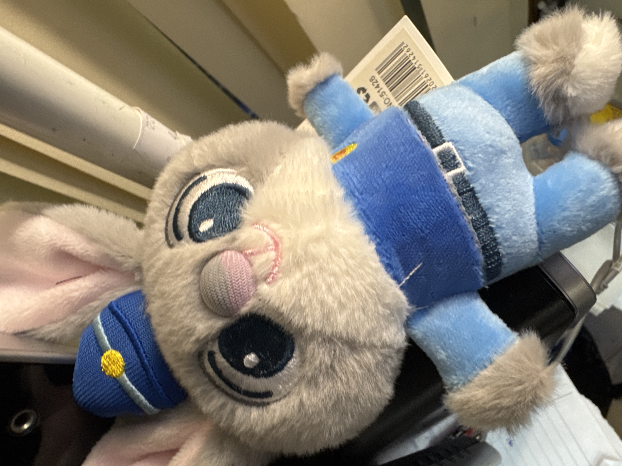 This close-up image features a plush toy rabbit, strongly resembling Judy Hopps from Disney's Zootopia, dressed in a police uniform. The toy is light gray and white with pink inner ears and a textured pink nose. Its large, round eyes are embroidered in dark blue with white highlights, giving it a stylized, cheerful expression. A small blue police cap with a yellow embroidered button and a light blue stripe is worn on its head. The uniform consists of a dark blue top with lighter blue sleeves and pants, accented with a darker blue belt and fuzzy light brown cuffs on the sleeves and at the bottom of the legs.
The toy appears to be resting on an unknown surface in an indoor setting. In the background, out of focus, are various objects, including white cylindrical items (possibly rolled papers or tubes) and a dark, indistinct object. A white tag with a barcode is visible behind the toy's shoulder. Visible text on the tag includes "NO-51428," the numbers "261514262" as part of the barcode, and the partial word "SON." The lighting suggests an artificial indoor light source. No activity is taking place; the toy is static.