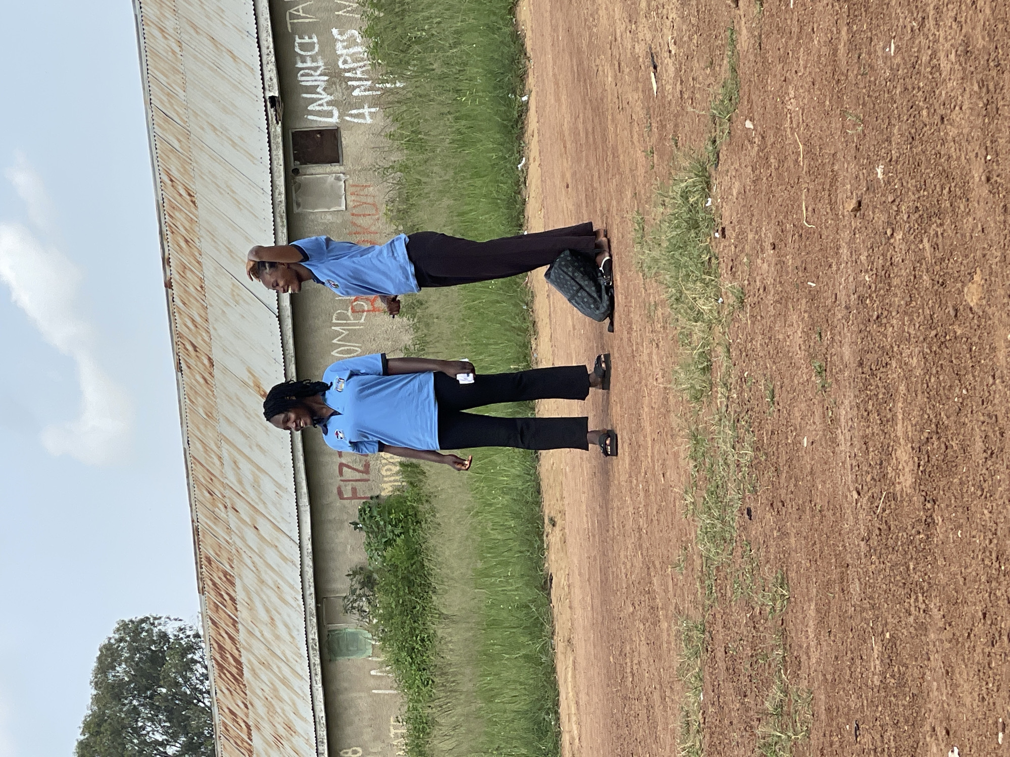 Two women are standing in a dirt lot in front of a long, corrugated metal-roofed building in Durumi, Nigeria. The woman on the left, with braided hair, wears a light blue polo shirt and black pants. She is looking to her left and smiling. The woman on the right, with lighter colored hair, also wears a light blue polo shirt and black pants. She has her right hand raised to her head, and she is looking to her left and smiling. A dark backpack is on the ground between her feet. The building behind them is weathered, with a light-colored facade that has graffiti on it. The ground is reddish-brown dirt with sparse green grass growing in patches. The sky is overcast.