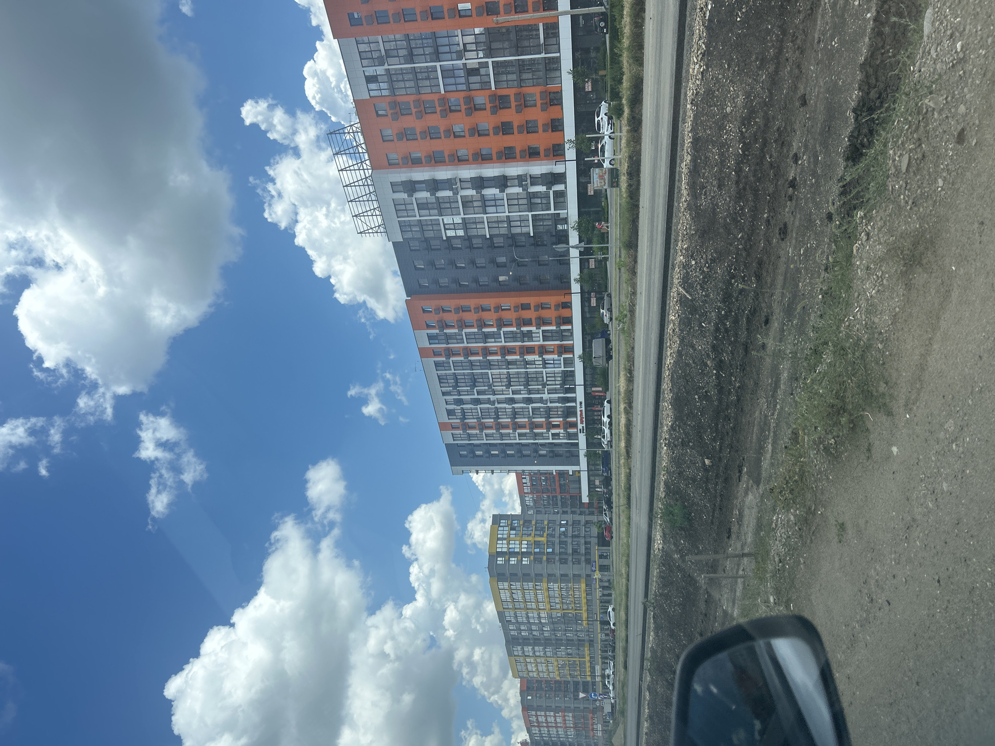 The image depicts a street scene in Orlovskii, Russia, featuring a row of modern, multi-story apartment buildings. The buildings have a varied color palette, with sections in orange, gray, and white. Numerous windows are visible on each building.
The scene appears to be set during the daytime, under a bright blue sky dotted with fluffy white clouds. The weather seems fair and sunny.
To the right of the buildings, there is a road, and beyond that, a strip of undeveloped land with gravel, dirt, and sparse vegetation. Several cars are parked along the street in front of the buildings. One car is a white SUV, and other vehicles are less distinct. There are also streetlights and some signage visible.
The perspective suggests the photo was taken from a vehicle, possibly through a side window or from the driver's seat, as a portion of a car's side mirror is visible in the foreground. The image is oriented vertically, with the sky occupying the left side and the buildings and road on the right. No people are clearly discernible in the frame, although the presence of cars and buildings implies human habitation. The overall impression is one of a residential urban or suburban area on a clear day.