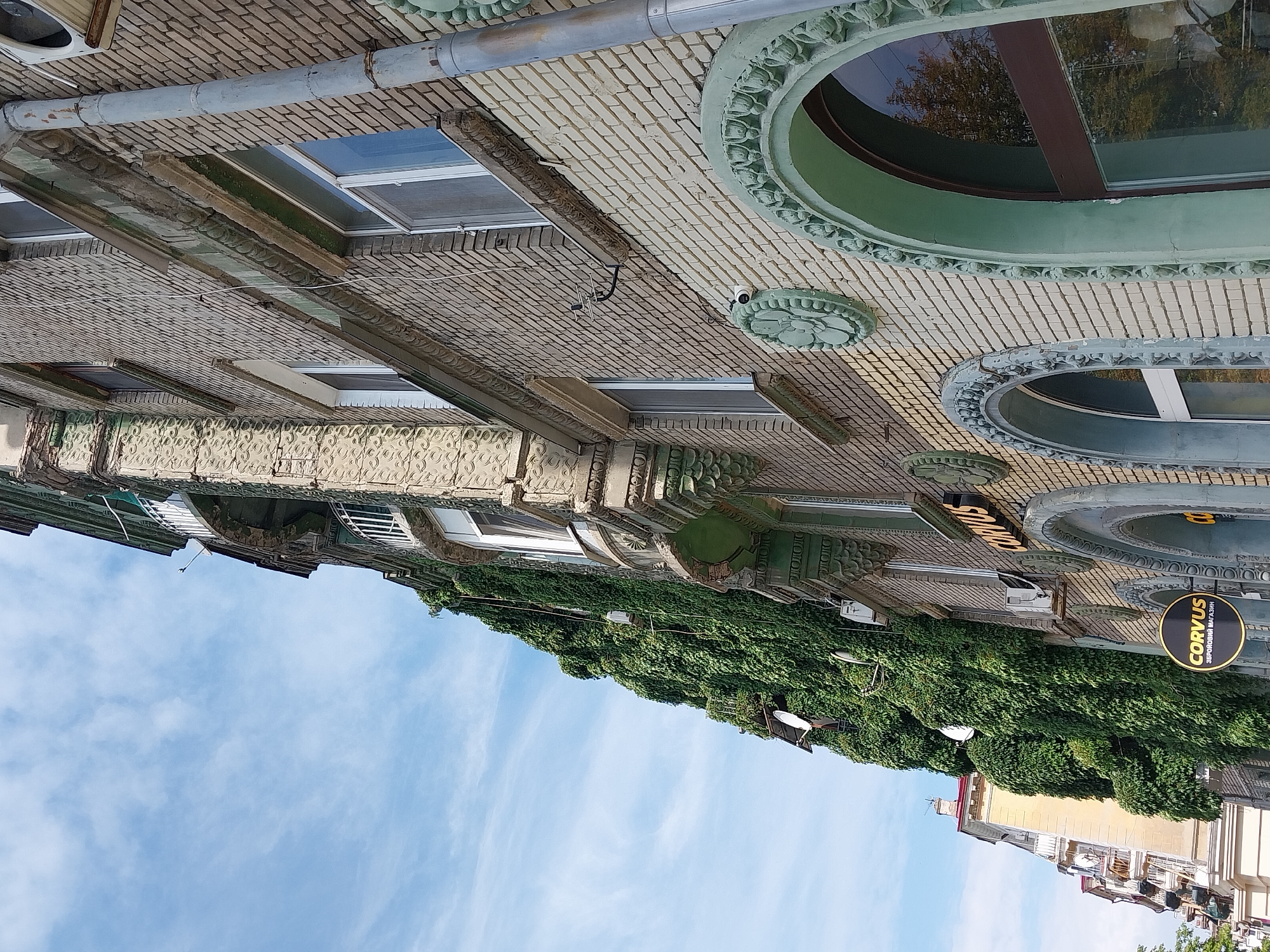 The image depicts a close-up, low-angle view of the exterior of a multi-story building in Dnipro, Ukraine. The building features brickwork of varying colors, with a prominent section covered in dense green ivy, creating a striking contrast. The architecture shows ornate detailing around windows and along the facade, with decorative stucco elements and arched window frames, some painted in a greenish hue. Several windows are visible, some with white frames and others with darker frames reflecting the surroundings. Air conditioning units and satellite dishes are mounted on the exterior wall. Two signs for "CORVUS" are visible, one larger illuminated sign and a smaller circular sign below it that reads "CORVUS ЗБОРОВИЙ МАГАЗИН," indicating a shop. The sky above is blue with wispy white clouds. To the left, another building with yellow facade and balconies is partially visible, suggesting an urban setting. The overall impression is one of an old, perhaps historical, building with a modern commercial establishment.