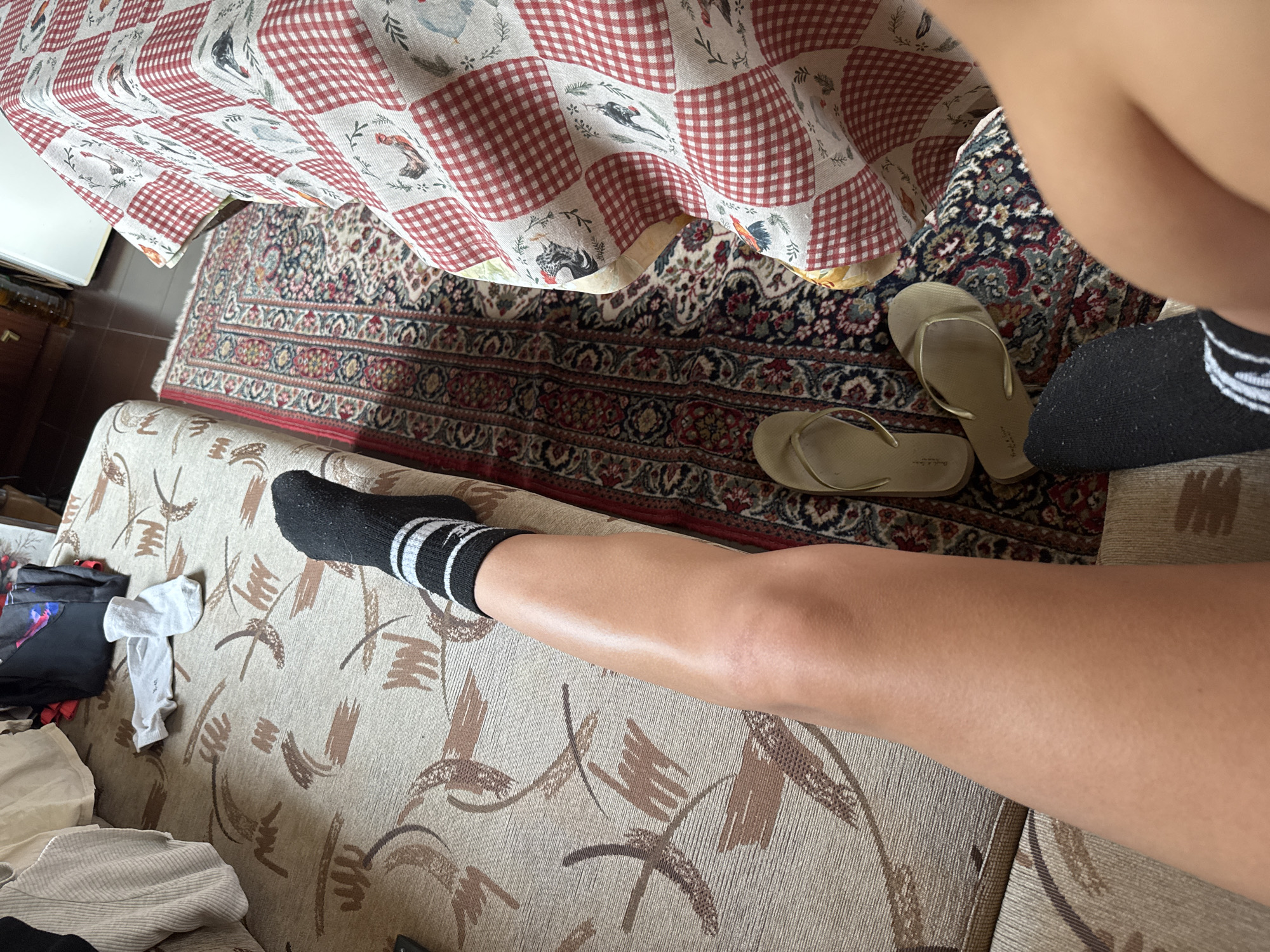 The image shows a portion of an interior space, likely a living room or similar domestic setting, in Drenovets, Bulgaria. A tan and brown patterned couch takes up much of the lower portion of the frame. Scattered on the couch are articles of clothing, including socks (one appears to be black with white stripes) and other miscellaneous items. A person's leg, clad in a sock, is prominently positioned on the couch. Above the couch is a patterned rug with red, tan, and brown tones. Overhanging the rug is a red and white checkered tablecloth. A pair of tan flip-flops rests on the rug next to a black sock.