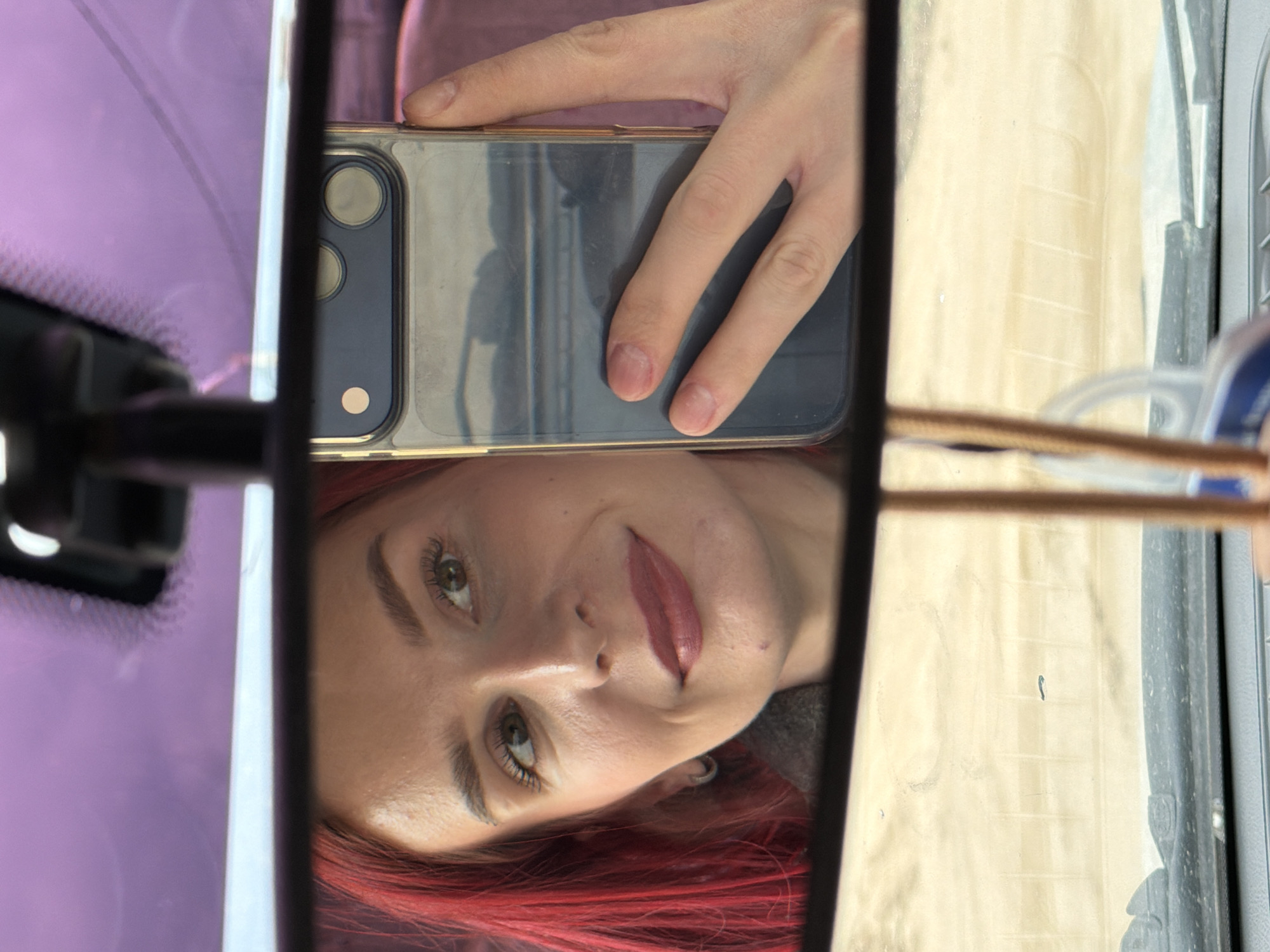 A person with red hair is taking a selfie in a car's rearview mirror. They are holding a phone with a clear case in front of the mirror. The reflection shows their face, hand, and the phone itself. The background visible through the windshield is blurred, with hints of light-colored structures. There is no definitive evidence in the image to identify Chaikovsky, Russia as the location.