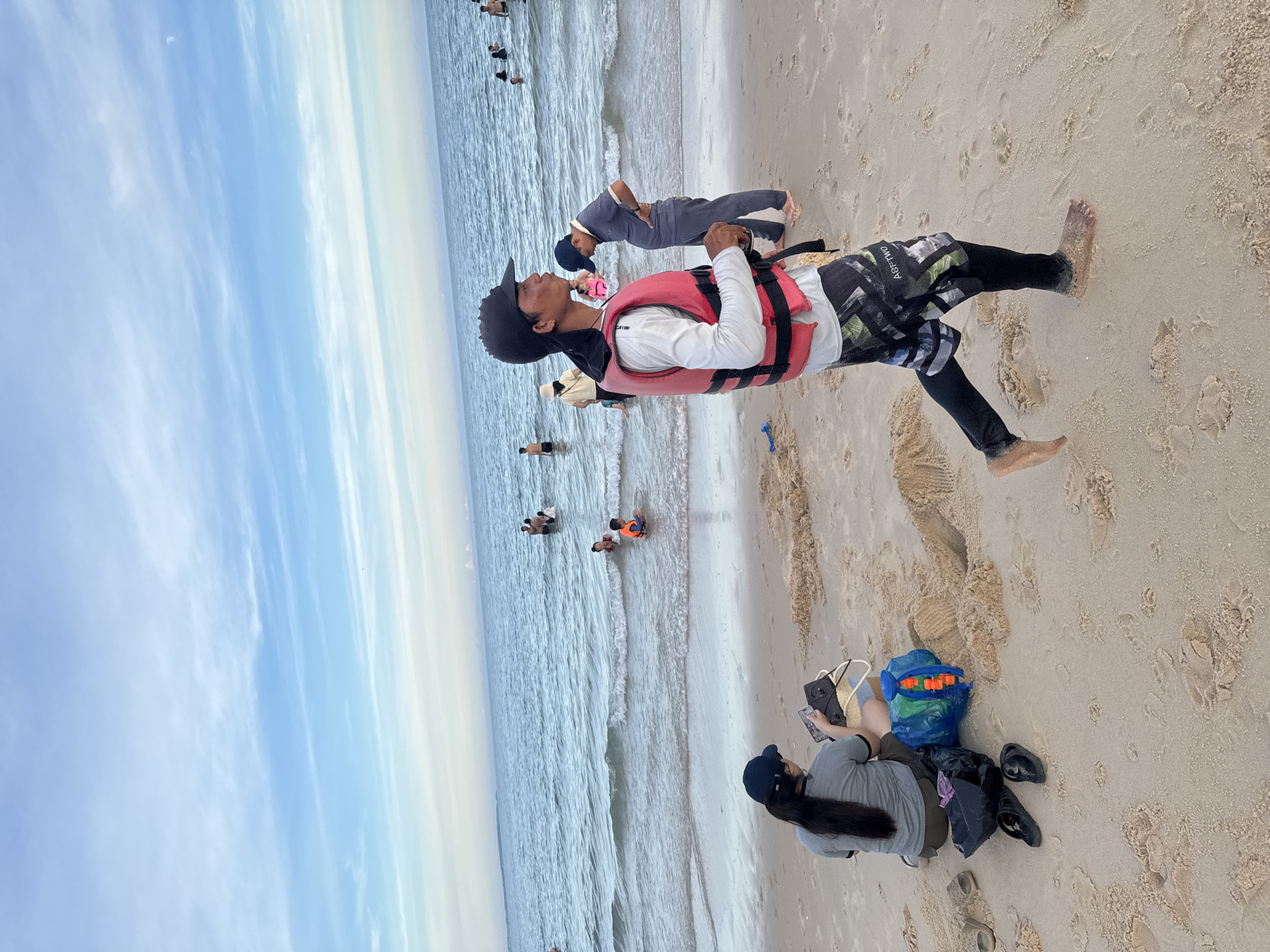 The image depicts a beach scene in Mũi Né, Vietnam. The primary focus is on a group of people enjoying the ocean and the sandy shore. In the foreground, a young man wearing a red life vest, a white long-sleeved shirt, and patterned shorts, stands on the wet sand, looking up. Beside him, on the sand, a person in a grey shirt and dark shorts is seated, holding a phone and a tablet. A blue bag with orange toys and a black trash bag are also visible near the seated person.
In the background, several other individuals are in the shallow water. One person is lying on their back in the water, while others are standing or wading. The sea is calm with gentle waves breaking on the shore. The beach is sandy, with footprints visible. The sky is a mix of blue and white, suggesting it is daytime with some clouds.
The overall atmosphere appears relaxed and recreational. The presence of a life vest and the activity in the water suggest swimming or water sports. The location cues, such as the style of the beach and the general appearance of the people, are consistent with a Southeast Asian coastal resort. No specific time of day can be definitively determined, but the lighting indicates it is likely daytime. The weather appears pleasant for beach activities.
The word "ASFFTIVO" is visible on the shorts of the man in the foreground.