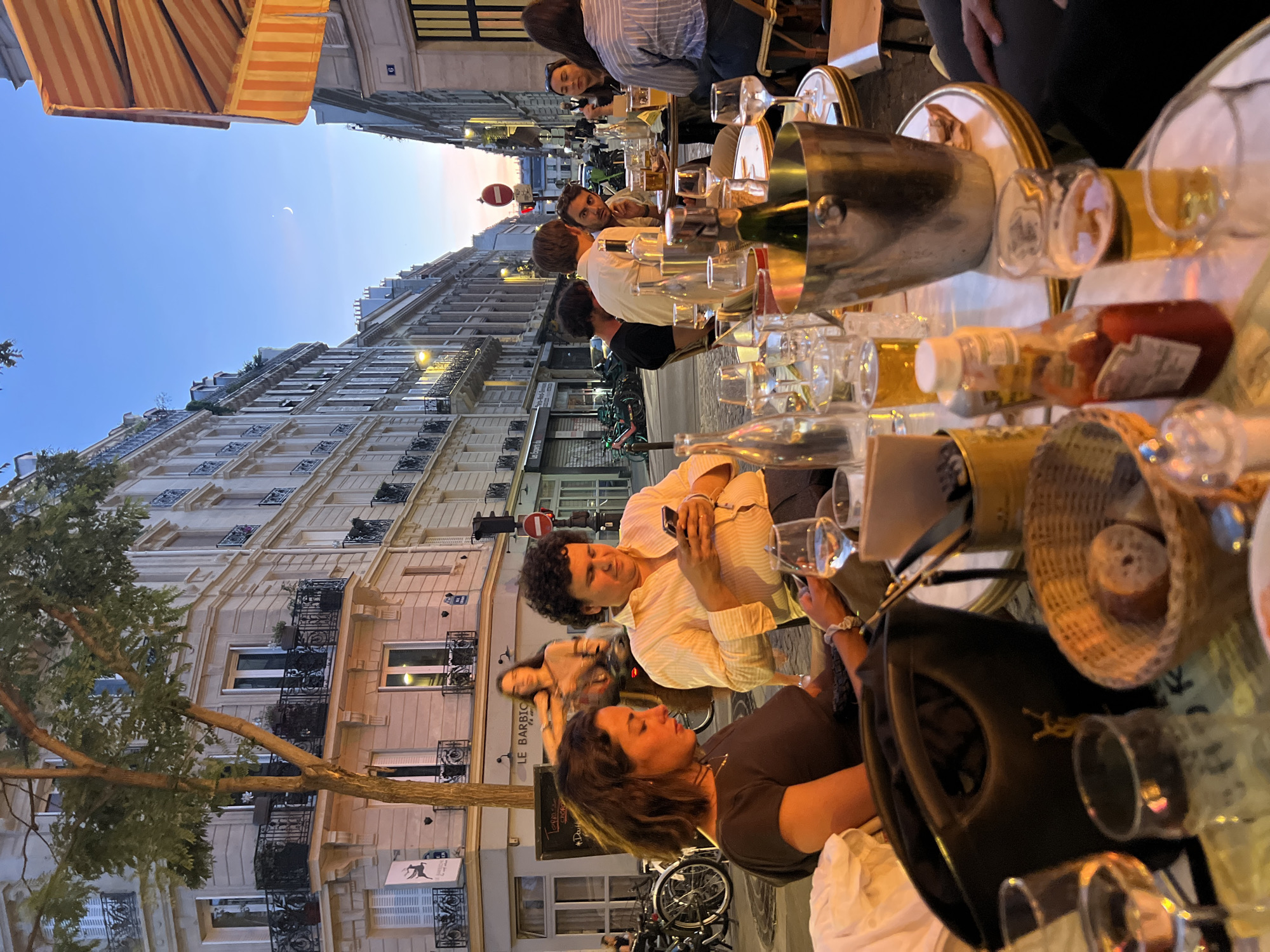 This image depicts an outdoor dining scene at a cafe in Paris, France, as dusk settles. The architecture of the buildings suggests a Haussmannian style, characteristic of the city. In the foreground, a table is laden with drinks, including wine glasses and beer. A black handbag with a gold logo is prominently placed on the table. To the right, a person in a striped shirt is seated, holding a phone. In the mid-ground, a man with curly hair is also looking at his phone, seated next to a woman. Behind them, a woman on a bicycle is visible, as is a sign that reads "LE BARBICHE". The street is lined with buildings and lampposts, and the sky is a deep blue with a sliver of moon visible. The overall atmosphere suggests a relaxed evening meal in a Parisian setting.