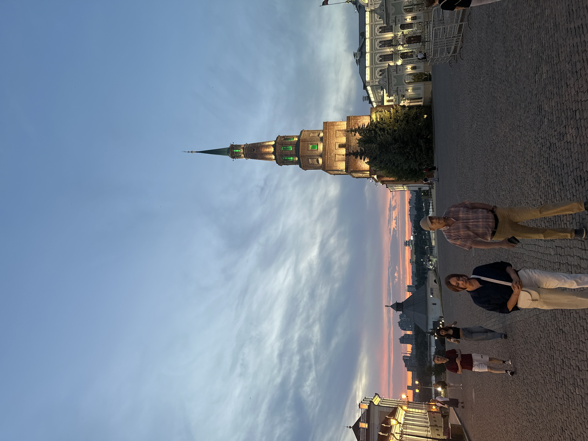 The image shows a view of the Soyembika Tower, located in the Kazan Kremlin in Kazan, Russia. The tower is the focal point, standing tall against a sky painted with hues of blue, gray, and pink from the sunset. People are standing or walking in a paved area near the tower. The building on the right has lighted windows.