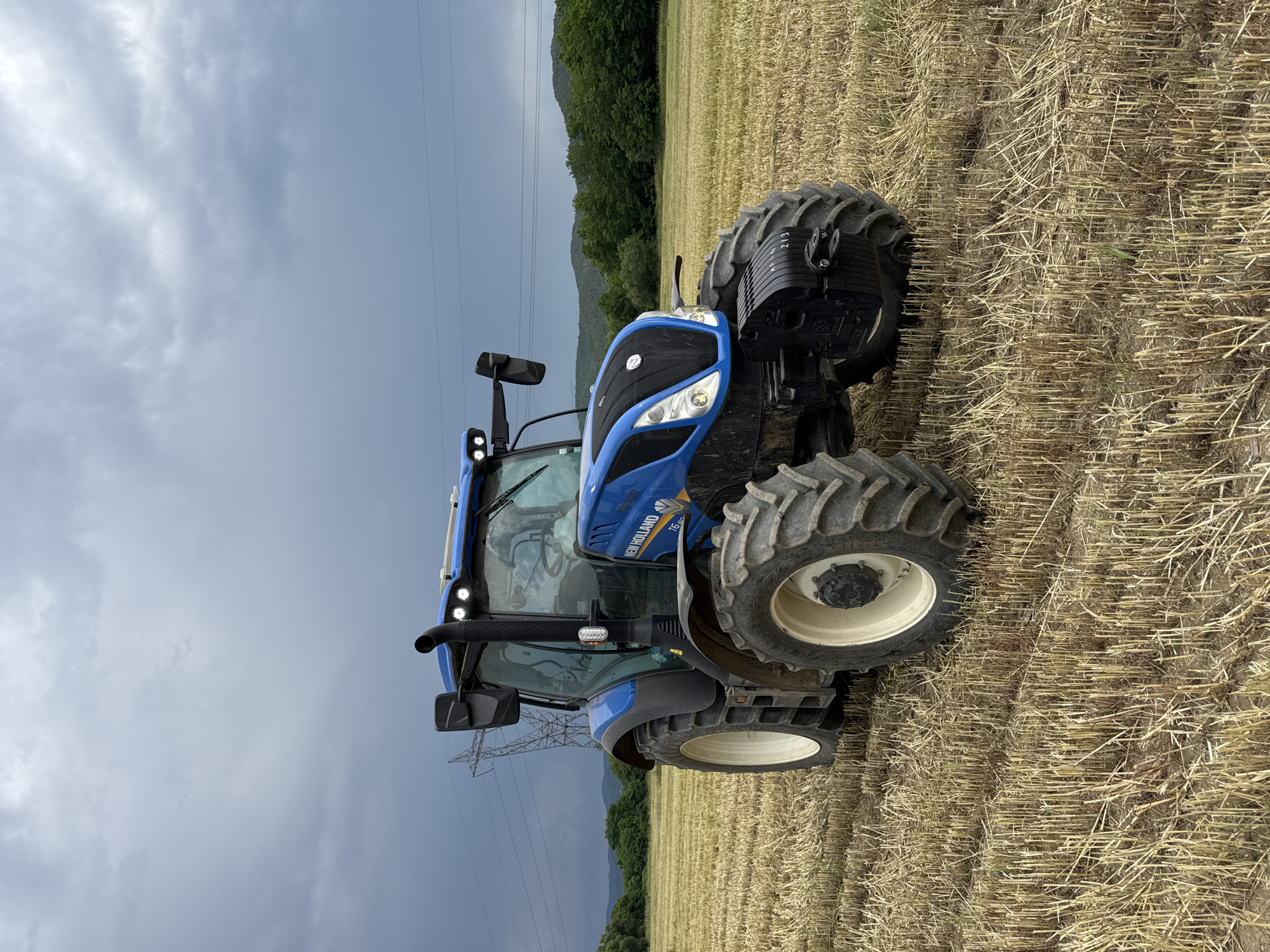 A blue New Holland T6.150 tractor is parked in a harvested field under a cloudy sky in Prolom, Bulgaria. The tractor is angled slightly to the left, with its front facing the right. The field is covered in dry, cut stalks of grain, indicating recent harvesting. In the background, rolling hills covered in green trees are visible, with a high-voltage power line tower and wires also present. The sky is overcast with dark, dramatic clouds. The tractor has large, rugged tires and a visible exhaust pipe. No people are present in the image.