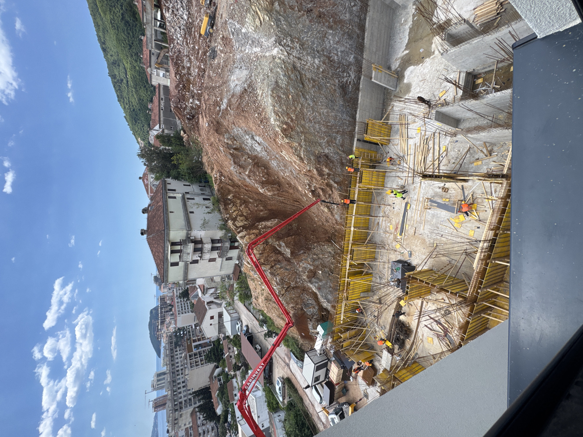 The image depicts a construction site in Budva, Montenegro. A large, reddish-brown rocky hillside dominates the center of the frame. Below, a construction crew is working on what appears to be the foundation of a building. Yellow wooden framework surrounds concrete structures, with several workers in orange vests and helmets visible. A red concrete pump truck is situated on the left side of the construction site. In the background, buildings of varying heights and styles, typical of an urban environment, rise towards the sky. A partially cloudy blue sky is seen above the buildings, indicating a sunny day.