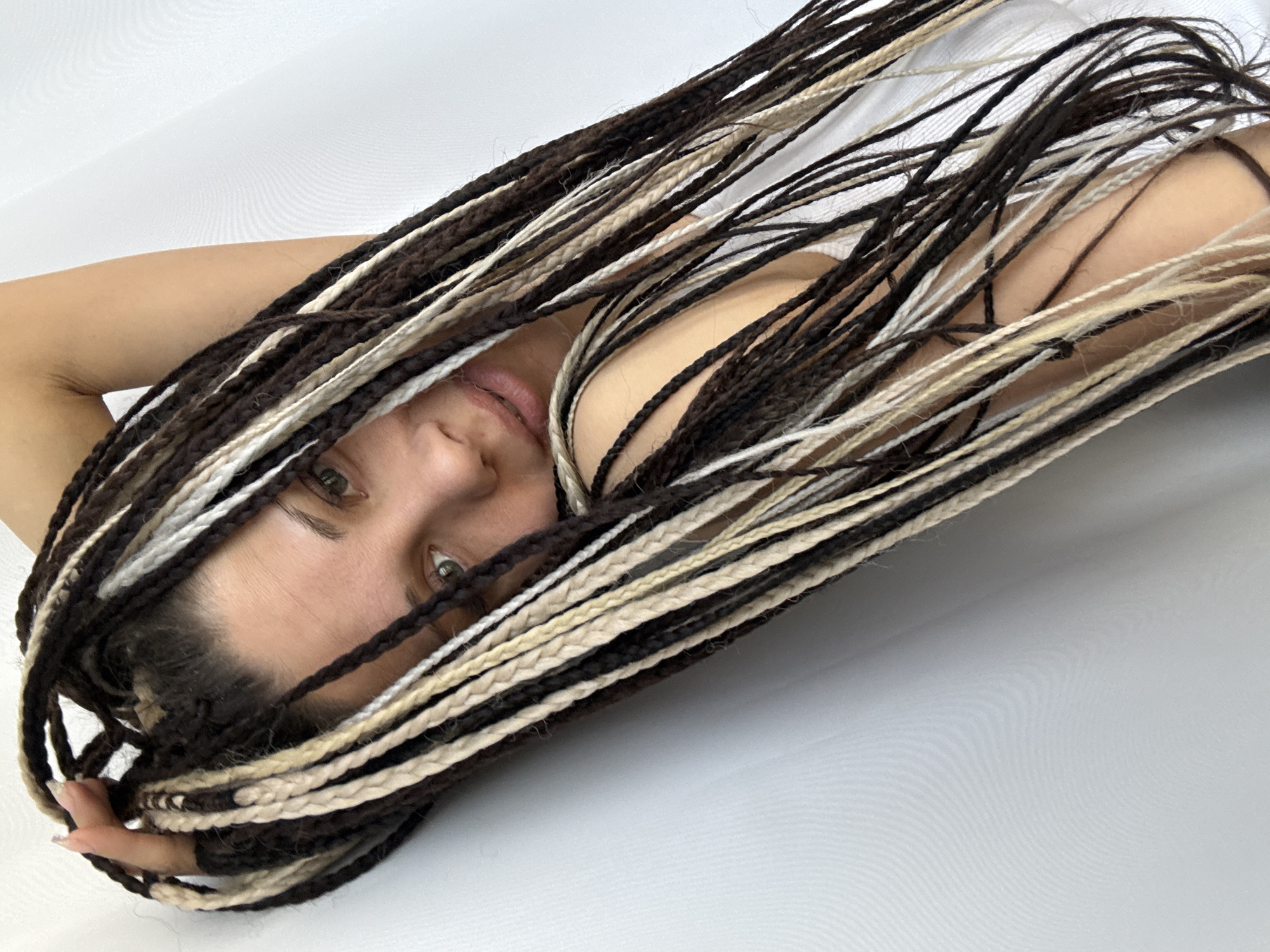 A woman with light skin and long, brown and blonde braided hair is lying on a white surface in Unknown, Unknown. Her head is tilted towards the camera, and her eyes are open. Her arm is visible on the right side of the frame. Some of the braids are obscuring her face.