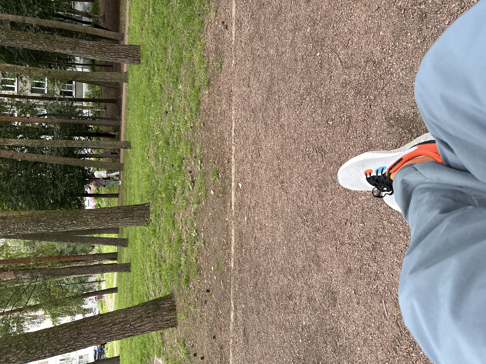 The image captures a view of a park in Vsevolozhsk, Russia, from a first-person perspective. The foreground shows the legs and feet of a person wearing gray pants and a white and orange athletic shoe. They are sitting on a gravel path, which is separated from a grassy area by a thin white line. In the background, a line of tall trees with rough bark stands on the edge of the grassy area. Beyond the trees, a multi-story residential building with white window frames is visible, partially obscured by the foliage. Two people are walking on a paved path in the middle ground, and a black car is parked to the left. The environment appears to be a public park with urban elements.