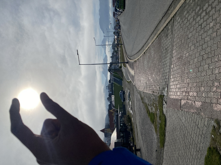 The image depicts an urban street scene in Ushuaia, Argentina, under a partly cloudy sky. In the upper left quadrant, a human hand with a visible blue sleeve is silhouetted against the bright sky, positioned to optically "pinch" or "hold" the sun, which appears as a brilliant white disc.
The foreground features a paved sidewalk composed of interlocking gray-red pavers, partially wet, alongside an unpaved gravel area with patches of grass and puddles. A curved asphalt road extends from the right foreground, bordered by a yellow line. Numerous dark-poled streetlights with rectangular fixtures line the road, where a queue of vehicles, including a green van, is visible on the right.
Further left, a cluster of buildings is present, notably one with a red-tiled roof and a steeple. A green and gray pedestrian overpass with stairs spans an area behind these structures, near which a white SUV is parked. The background reveals rugged mountain ranges, partially obscured by dense, low-hanging clouds or fog. The sky above is predominantly covered by stratocumulus clouds, with the sun breaking through centrally. The scene is illuminated by daylight.