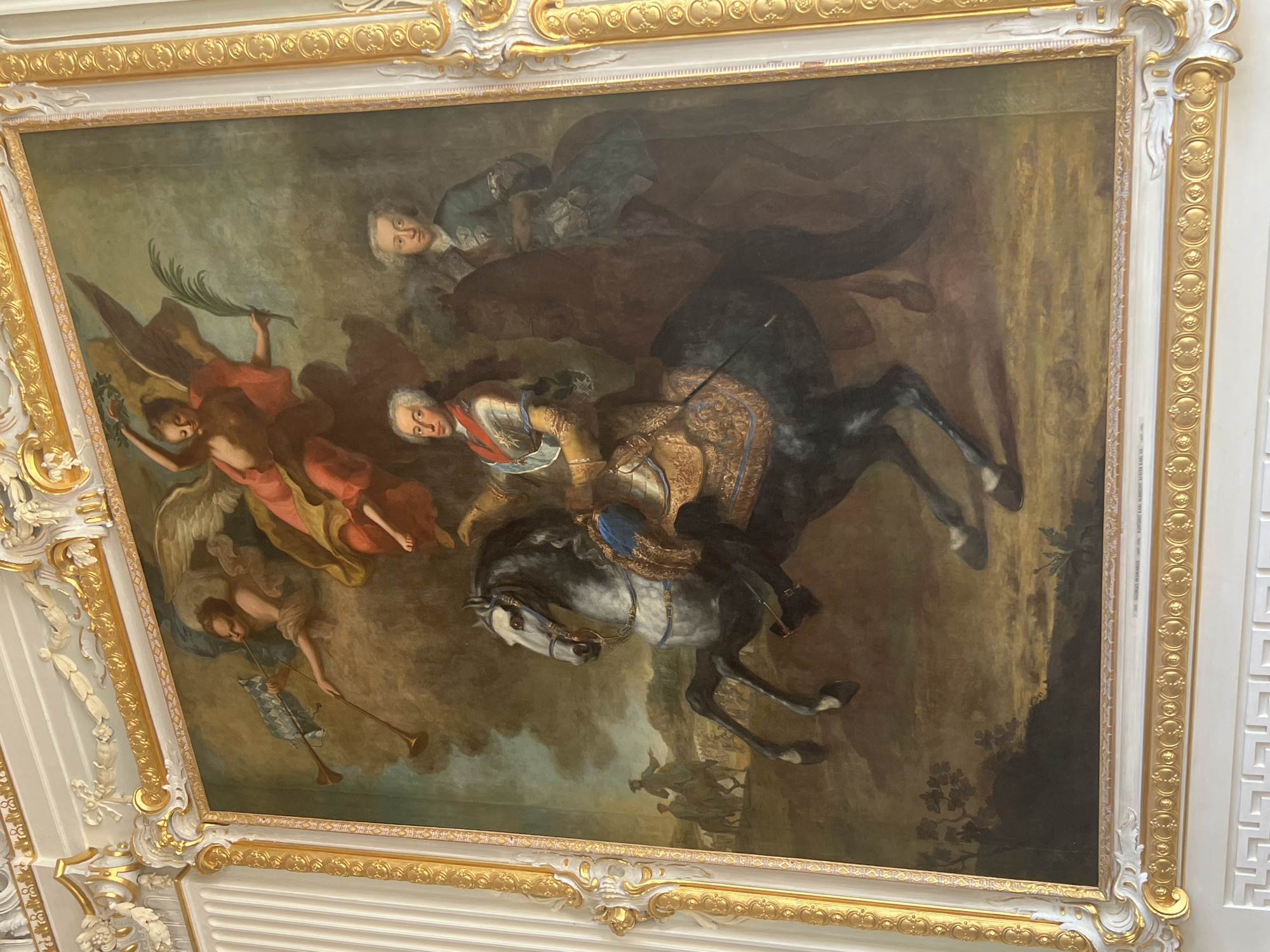 This is a full-length oil painting of a man on horseback in a formal setting, likely a portrait of a historical figure. The painting is housed in a richly ornate gold and white frame. The central subject is a man in elaborate military attire, including armor and a sash, riding a grey horse with a dark mane. He holds the reins in his left hand and appears to be looking forward.
Above and behind the central rider, two winged figures are depicted in the clouds. One figure, with red drapery, holds a laurel wreath and a palm frond. Another figure, with lighter drapery, plays a trumpet, and a checkered flag is visible behind the trumpet. To the right of the central rider, another man on a darker horse is visible, wearing a blue coat and white wig. Further in the background, on the left side, two figures on horseback are seen in a landscape with a group of soldiers.
The painting is situated in the **Neues Schloss (New Palace)** in Oberschleißheim, Germany. The opulent frame and the grand subject matter suggest it is a significant artwork within the palace's collection.