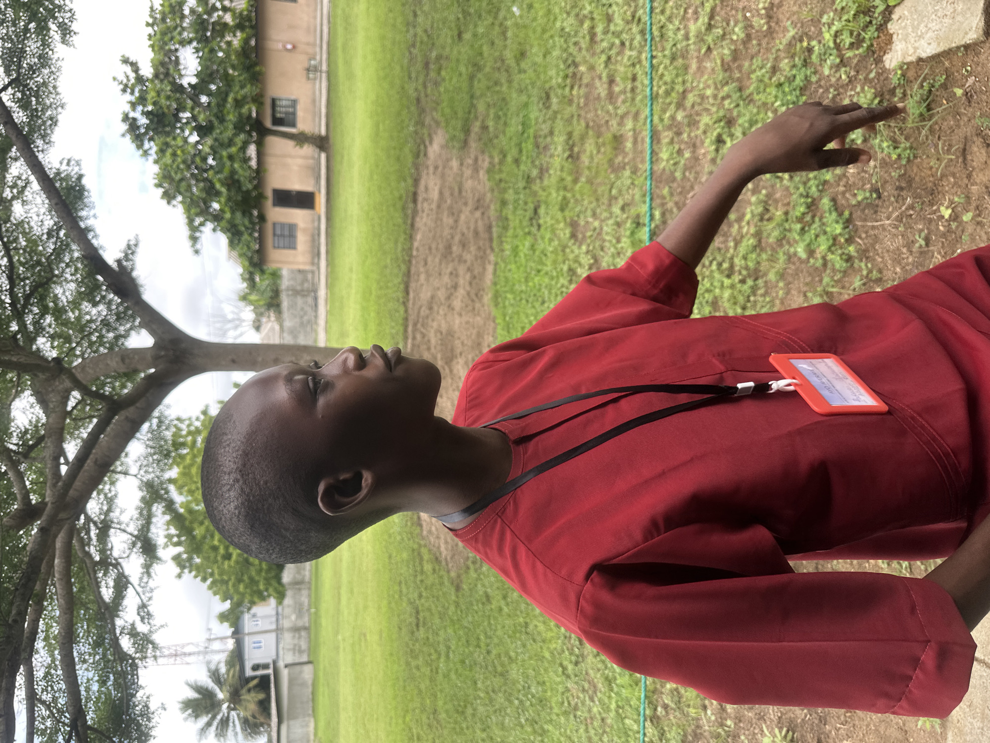 A young person with dark skin and a short haircut is shown from the chest up, facing to the right. They are wearing a red tunic with short sleeves and a black lanyard with an orange ID badge attached. The background is an outdoor scene with a large tree overhead, a grassy field, and a light brown building with windows and a door. The building appears to be a single-story structure with a corrugated roof. In the distance, there are other trees and a palm tree. A cell tower is visible on the left side of the image. The lighting suggests it is daytime, possibly overcast. The overall setting is rural or suburban.