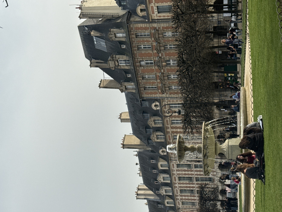 The image depicts a public square identified as Place des Vosges in Paris, France, under an overcast sky. In the immediate foreground, a patch of vibrant green grass occupies the lower third of the frame. Four young individuals are seated directly on this grass in the lower-left corner; three face away or towards each other, while one in a red top faces right. They appear to be engaged in conversation or resting.
Mid-ground, a multi-tiered, ornate stone fountain actively streams water into its basins. Surrounding the fountain and extending towards the background are numerous bare deciduous trees, their branches devoid of leaves, suggesting a cold season. Beyond the grass and fountain, a paved area with visible pathways and low decorative fencing is populated by several individuals walking or seated on benches beneath the trees. A distinctive black wrought-iron lamppost with a spiral design stands to the right of the fountain.
The background is dominated by a continuous row of historic Parisian buildings characteristic of Place des Vosges. These structures feature multiple stories with red brick facades, highlighted by white stone quoins and window surrounds. They are topped with steep, dark gray Mansard roofs, punctuated by numerous dormer windows of varying designs and multiple tall, rectangular chimneys. A modern-looking, flat, reflective panel is visible on the roof of one building towards the right side of the frame. Archways are discernible at ground level behind the trees, forming a continuous arcade along the building base.