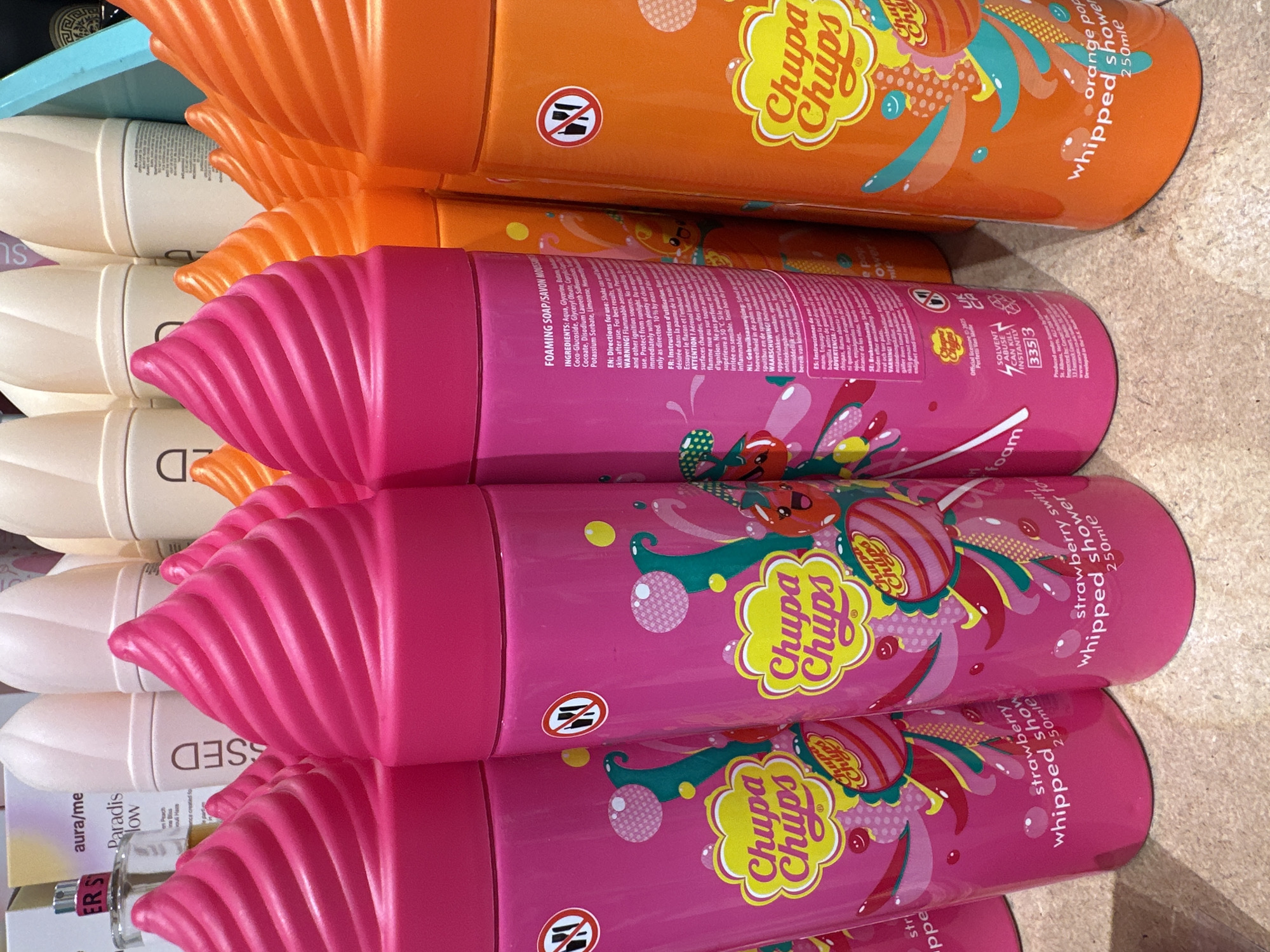 In an indoor retail environment, a close-up, eye-level shot captures a collection of vibrantly colored body wash bottles arranged on a textured, light-brown surface. The bottles are designed to resemble ice cream cones, with swirling tops in bright pink and orange. The pink bottles feature the "Chupa Chups" logo and a strawberry-themed design with cartoonish fruit characters and bubbles. The orange bottles also bear the "Chupa Chups" logo and appear to have an orange soda or fruit-themed design. In the background, several neutral-colored aerosol cans and the edge of a blue display are visible. The lighting suggests an artificial retail setting.