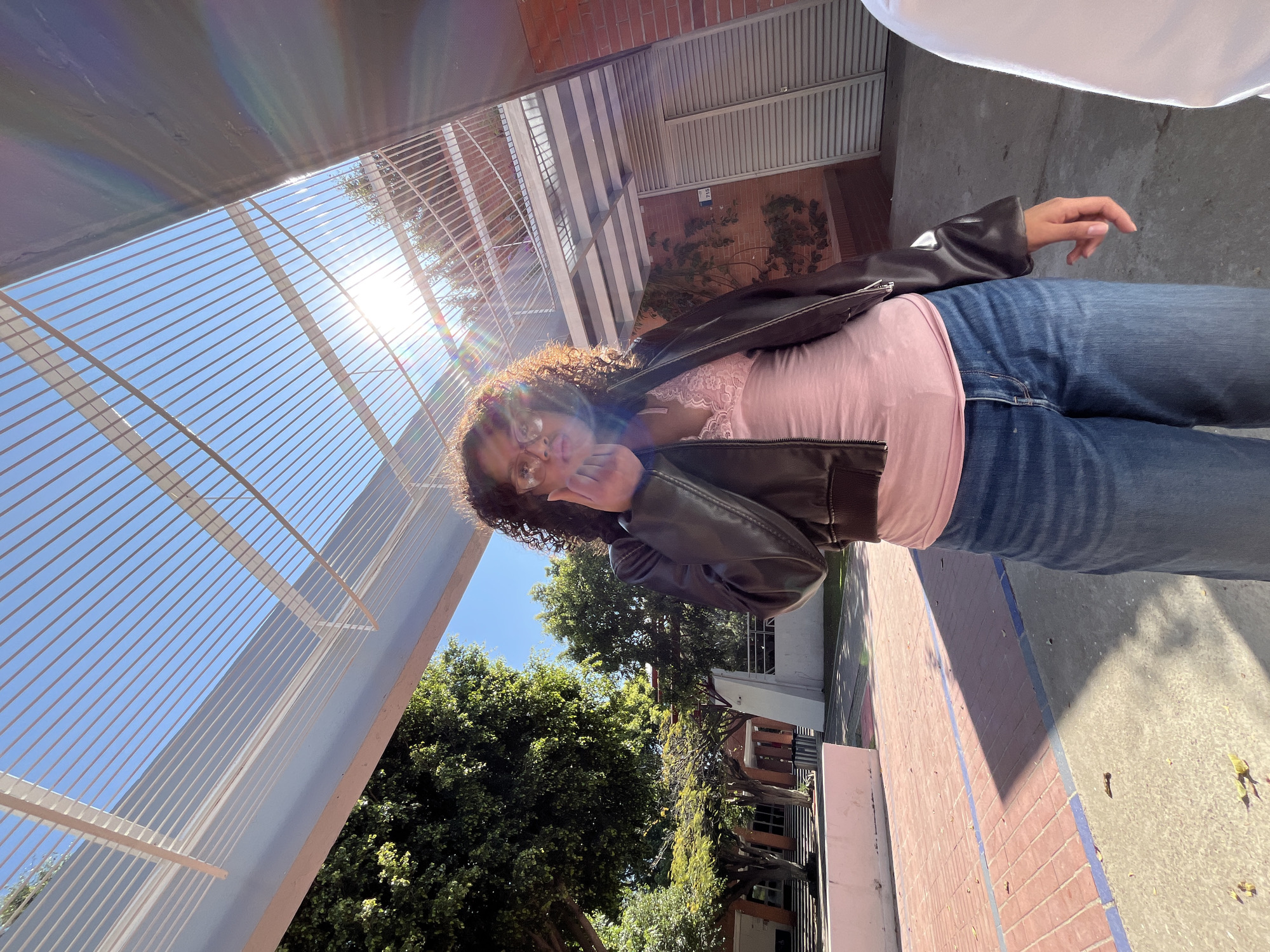 An outdoor scene depicts a young woman standing centrally under a large, light-colored metal pergola structure with numerous parallel horizontal bars. She has curly dark hair, wears glasses, a dark brown/black leather-like jacket over a pink lace-trimmed top, and blue jeans. Her right hand is raised near her mouth, and she looks directly at the camera. The ground is a mix of concrete and red brick paving with blue demarcation lines, scattered with a few fallen leaves. Strong sunlight from the upper right creates lens flare and distinct shadows on the ground. To the left, several large green trees are visible, along with a concrete planter/bench and the facade of a multi-story building featuring stairs. To the right, a brick wall is visible with climbing vines, alongside a gray louvered/slatted door or shutter marked with the number "715." A white garment, belonging to another partially visible individual, is present on the far right edge of the frame. The location is the campus of the Universidad de las Américas Puebla (UDLAP) in San Andrés Cholula, Puebla, Mexico.