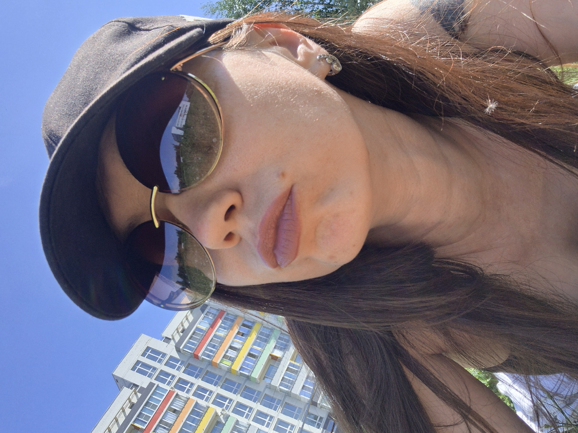 A low-angle, tilted, medium-shot photograph captures a light-skinned woman with brown hair. She wears a dark-colored baseball cap and sunglasses with gold-colored frames. The woman is looking down at the camera.
In the background, there is a partial view of a multi-story building. The building is predominantly white with rectangular windows. Some of the windows are highlighted with pastel colors. The sky is blue.