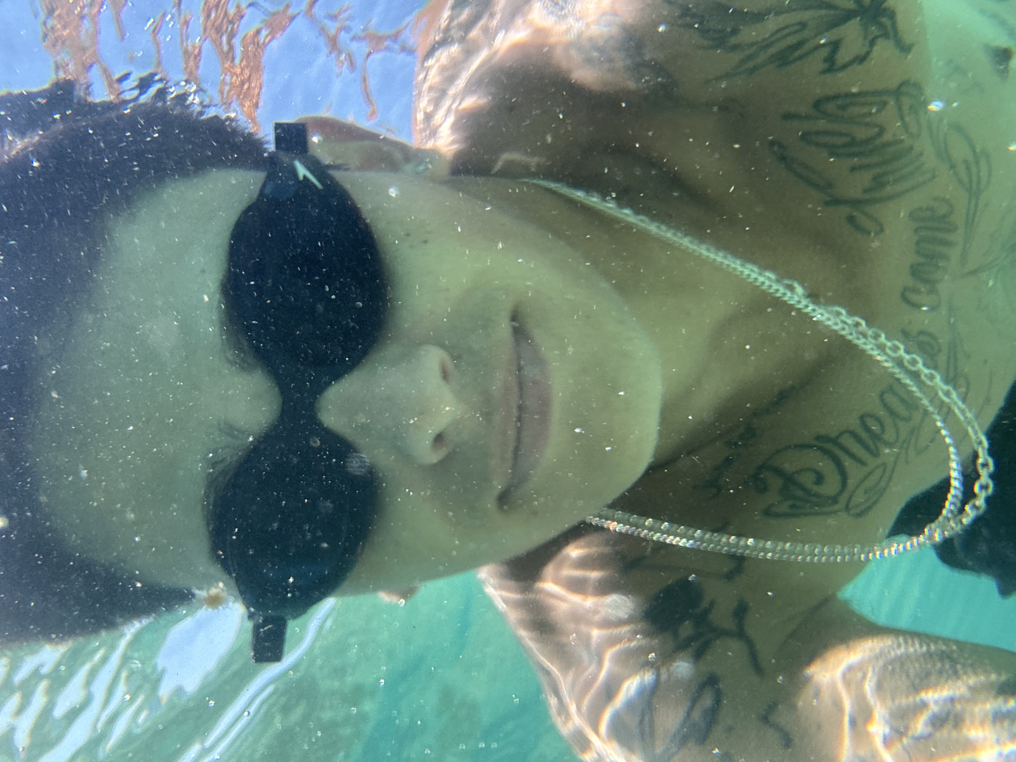An underwater selfie is taken by a young person wearing black swimming goggles. They have dark hair and visible tattoos on their chest and arms, including the words "Dream" and "true" on their chest. A silver chain is worn around their neck. The water is turquoise and clear, with some particles and light refractions visible. In the background, there are hints of seaweed and the surface of the water. The scene appears to be taken in a natural body of water, possibly the Mediterranean Sea off the coast of Arenys de Mar, Spain, given the clear turquoise water and presence of seaweed.