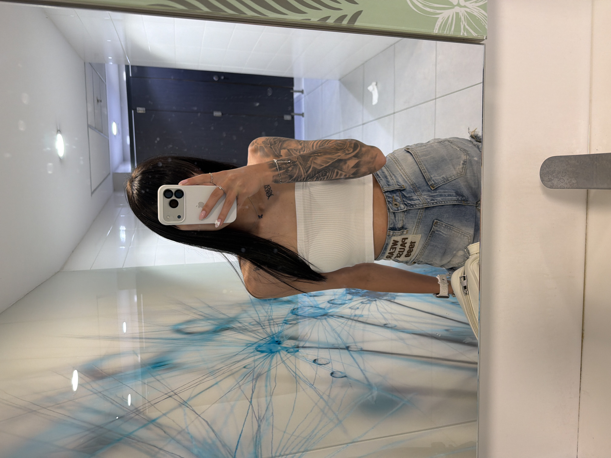 A young woman is captured in a mirror selfie within a public restroom at Plovdiv Airport, Bulgaria. She stands, reflected in a large rectangular mirror, with long, straight, dark hair partially obscuring her face. She wears a white ribbed strapless crop top, light blue distressed denim shorts with a visible label (reversed "RAW PAUSE WEAR REER"), and a white watch on her left wrist. Her left arm is adorned with a full sleeve tattoo, and a smaller dark tattoo is present on her upper chest. Her right hand, featuring light pink fingernails with a white floral detail on one nail and multiple rings, holds a light-colored smartphone with a triple camera system, actively capturing the reflection. A white fanny pack is visible on her left hip. The environment behind her, reflected in the mirror, includes a distinct white wall with an abstract blue mural featuring radiating lines and circular elements on the left. To the right, white tiled walls reflect a dark grey restroom stall door with metal hinges and handles. The floor is covered with white tiles, and a discarded white tissue is visible on it. Overhead, illuminated recessed ceiling lights are reflected in the mirror.