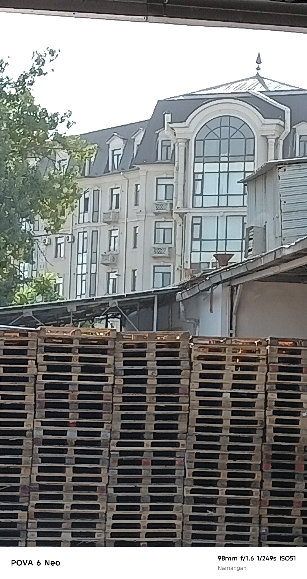 The image depicts a cityscape in Namangan, Uzbekistan. In the foreground, a large stack of wooden pallets is visible. Behind the pallets, a multi-story building with a grey, mansard roof and arched windows is the prominent architectural feature. The building appears to be in a state of construction or renovation, with some industrial elements, such as metal sheeting and a sloped roof structure, partially obscuring its facade. A tree with green foliage is visible on the left side of the image. The sky is overcast and bright. The provided text overlays indicate the camera model and settings: "POVA 6 Neo" and "98mm f/1.6 1/249s ISO51", along with the location "Namangan".