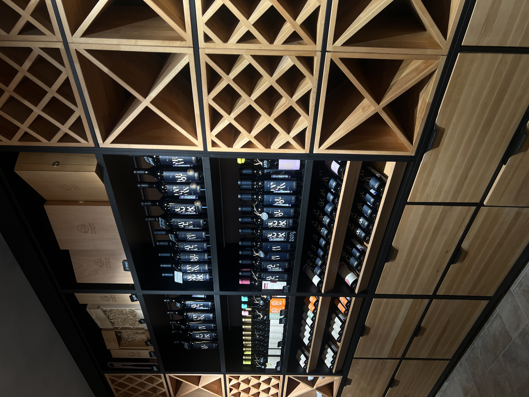 An interior view depicts a modern wine storage and display area, likely a retail space or cellar, located in Vinogradnoe, Russia. The scene is dominated by floor-to-ceiling shelving units crafted from light-colored wood and black metal frames.
On the left and right, extensive wooden wine racks feature a prominent diagonal grid pattern, forming numerous square and triangular compartments. These racks are largely empty in the upper sections but contain some horizontally stored bottles in the lower parts.
The central section comprises multiple black shelves displaying an organized collection of wine bottles. The top two shelves prominently feature dark-colored bottles, predominantly with black labels bearing white or light-colored script and the "УСАДЬБА МАРКОТ" (Usadba Markot) logo. Price tags are visible on many bottles (e.g., 5500, 3600, 2650, 3100 Rubles). Above these bottles, light-colored wooden gift boxes are stacked, some closed and some open, revealing straw packing material, also bearing the "УСАДЬБА МАРКОТ" emblem.
The lower central shelves present a mix of wine bottles, including some with lighter-colored labels (white, pink, light green, orange) standing upright. Below these, additional rows of dark-labeled bottles are laid horizontally in wooden cradles.
The entire lower portion of the shelving system consists of flush, light-colored wooden drawers with minimalist black horizontal handles. The floor is covered with large, square, light brown or greyish tiles. No individuals are present in the scene, and no active interactions are depicted. The lighting is focused, creating some shadowing while highlighting the displayed merchandise.