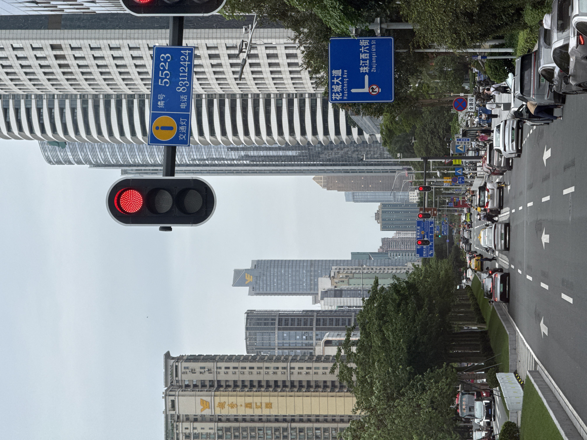 The image shows a street scene in Tianhe District, Guangzhou, China. A row of cars is stopped at a traffic light that is red. The street is lined with trees, and in the background are several high-rise buildings. The street signs indicate the location is at Huacheng Avenue (花城大道) and Zhujiangxi 6th Street (珠江西六街). There are also signs indicating street number 5523 and the phone number 83112424.