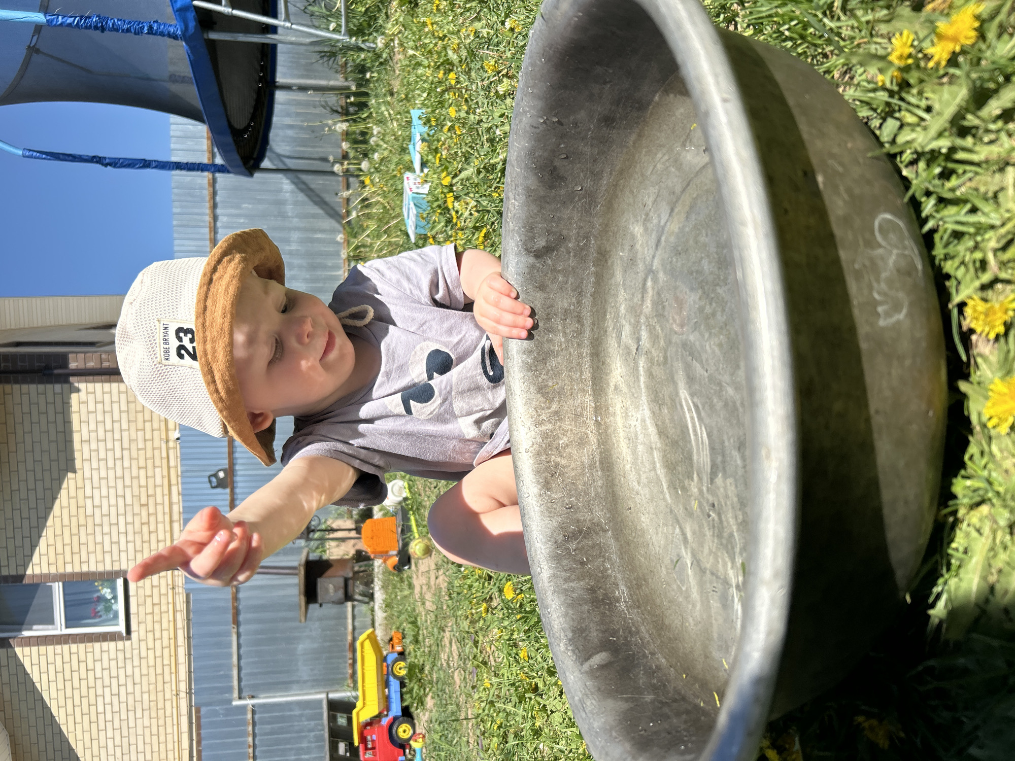 A young child is squatting in a grassy yard next to a large, metal washbasin. The child is wearing a gray t-shirt and a bucket hat. The hat has "KOBE BRYANT 23" printed on it. The child is pointing with their left hand toward the left side of the frame. Behind the child, there is a blue trampoline and a metallic fence. In the background, there is a brick building and some toys scattered on the grass. The grass is green and there are yellow dandelions growing in it. The photo was likely taken in the city of Chaikovsky, Russia.