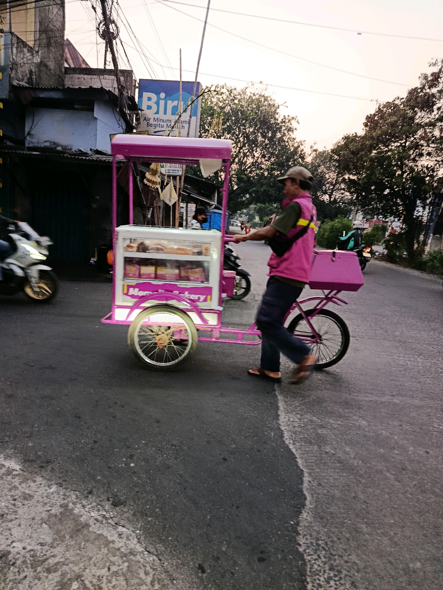 The image shows a street scene in the city of Bekasi, Indonesia. A man wearing a pink vest and a cap is pushing a pink food cart with a glass display case filled with bread. The cart is attached to a bicycle. A motorcycle is visible in motion on the left side of the image. In the background, there are buildings, trees, and overhead electrical wires. A blue sign with white lettering that says "Biru" and "Air Minum Alami Begitu Mujic" is visible on one of the buildings. The sky appears overcast, suggesting it might be late afternoon or early morning. The street is paved and appears to be a road in an urban or suburban area.