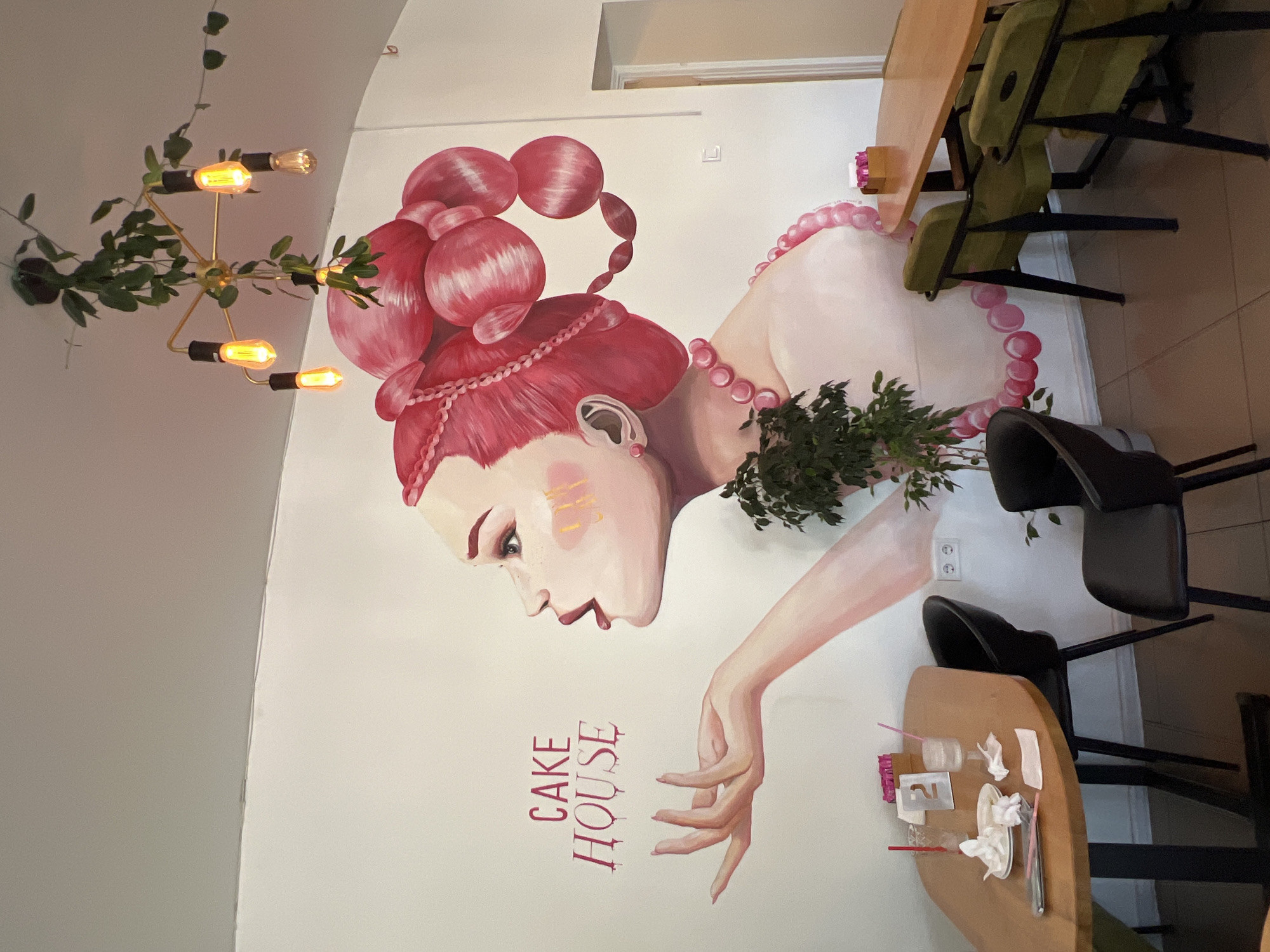 This image shows the interior of a cafe with a large mural on the wall. The mural depicts a woman with bright pink hair styled in elaborate buns, wearing a pink pearl necklace. Her profile is shown from the neck up, with her mouth slightly open and one hand extended. The text "CAKE HOUSE" is written in pink, dripping font to the left of the woman's head. Green plants are placed in front of the mural.
In the foreground, there are several tables and chairs. A wooden oval table is set with white napkins, glasses, and a small container with pink items. Black chairs are placed around this table. To the right, another wooden table is visible with green chairs. A modern chandelier with exposed bulbs and hanging greenery is suspended from the ceiling. The cafe appears to be located in Odesa, Ukraine, based on the provided context.