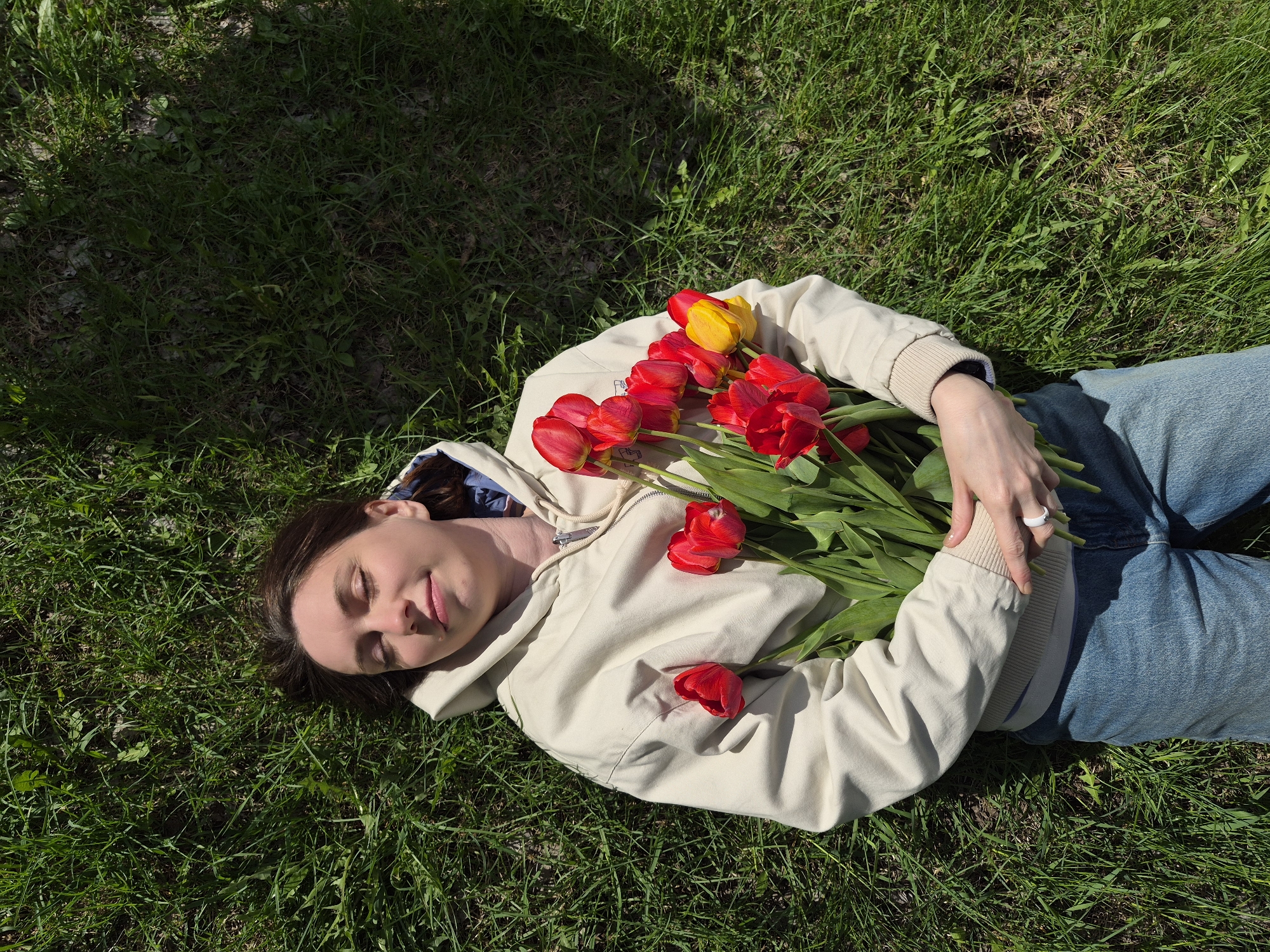 A woman lies on a patch of green grass, holding a bouquet of red tulips with one yellow tulip. The woman has brown hair and wears a light beige jacket and blue jeans. She has her eyes closed and appears relaxed. The setting is outdoors with sunlight and shadows on the grass. The location cannot be determined from the image.