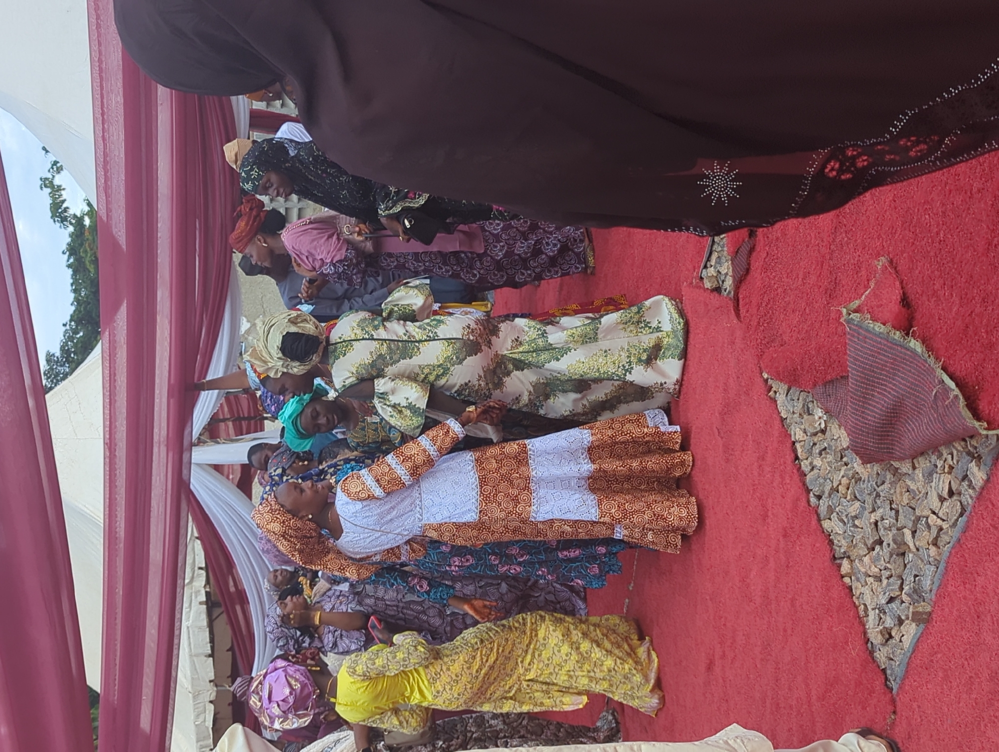 This image captures an outdoor social gathering in Kabala, Nigeria. The scene is illuminated by natural light under a partly cloudy sky, with white tents and draped maroon and white fabric forming an overhead canopy. The ground is covered with a bright red carpet, which has a triangular section cut out or missing in the foreground, revealing a bed of grey and light brown gravel. A crumpled piece of red, patterned fabric rests on the gravel.
Numerous individuals, predominantly women, are present, dressed in vibrant, patterned traditional African attire, including long dresses and elaborate headwraps (gele). In the center of the frame, two women appear to be interacting or dancing: one in a long yellow gown with a purple headwrap, and another in a white and orange patterned dress with an orange headwrap, facing each other. To their right, another woman wears a light-colored floral dress and matching headwrap. Other individuals, including men partially obscured, are visible in the midground and background, standing and conversing. In the immediate right foreground, the back and side of a person wearing a dark brown garment with intricate rhinestone or beaded embellishments on the sleeve are visible. The overall impression is a festive communal event.