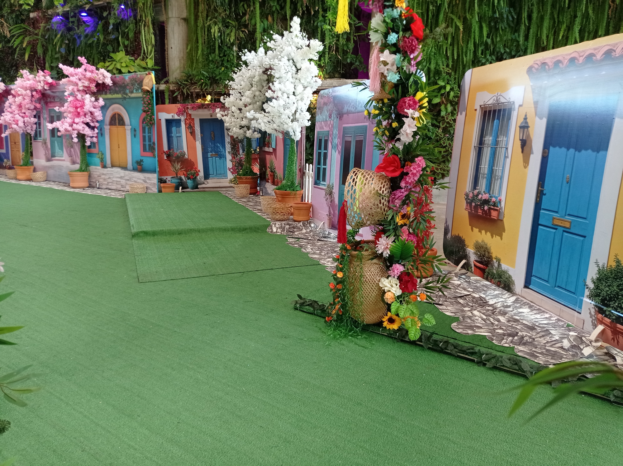 The image depicts a vibrant, artificial cityscape likely located in Pampatar, Venezuela, characterized by colorful facades reminiscent of traditional architecture. These facades are likely photo backdrops erected for decorative purposes or a staged event. The backdrops feature depictions of homes or shops with distinct architectural details: various painted doors, shuttered windows, and window boxes filled with flowers.
Artificial trees with white and pink blossoms are positioned in front of some of the facades to enhance the scene. The entire display is set on a green artificial turf, giving the impression of a miniature town square or pedestrian zone. The presence of woven baskets and floral arrangements further accentuates the scene. The shot appears to be taken from a slightly low angle. The scene is enclosed by a lush, green wall of plants in the background.