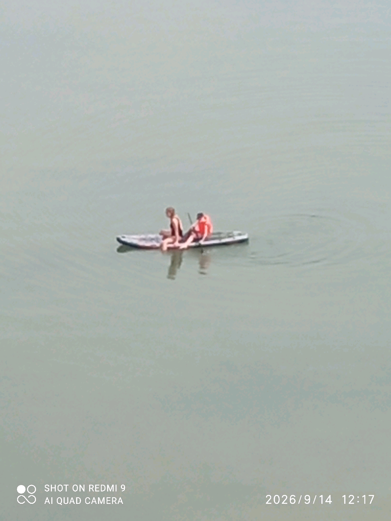 Two individuals are seated on a paddleboard in calm, light-colored water. The person on the left is wearing a dark top and appears to be looking forward. The person on the right is wearing an orange life vest and is looking to the left. The paddleboard is white with a patterned design on the bottom. The water has subtle ripples. The timestamp "2026/9/14 12:17" and "SHOT ON REDMI 9 AI QUAD CAMERA" are overlaid on the bottom of the image. The scene appears to be a recreational activity on a body of water.
