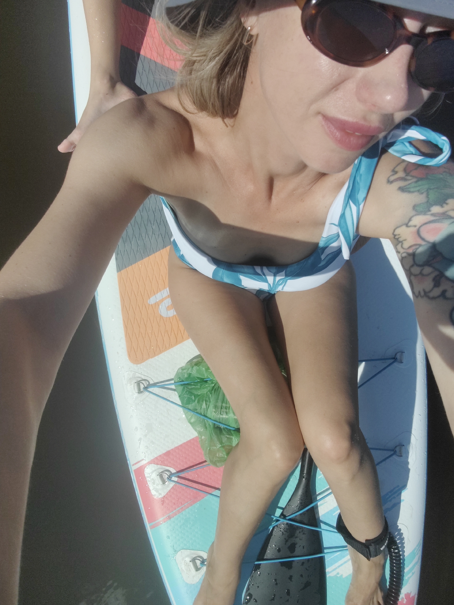The image shows a woman on a paddleboard in the city of Saint Petersburg, Russia. The woman is wearing sunglasses and a bikini, and she has tattoos on her arm. She is sitting on the paddleboard, and her legs are bent. A paddle is lying on the paddleboard next to her. The paddleboard is in the water, and the water is dark. The sun is shining, and the sky is blue.