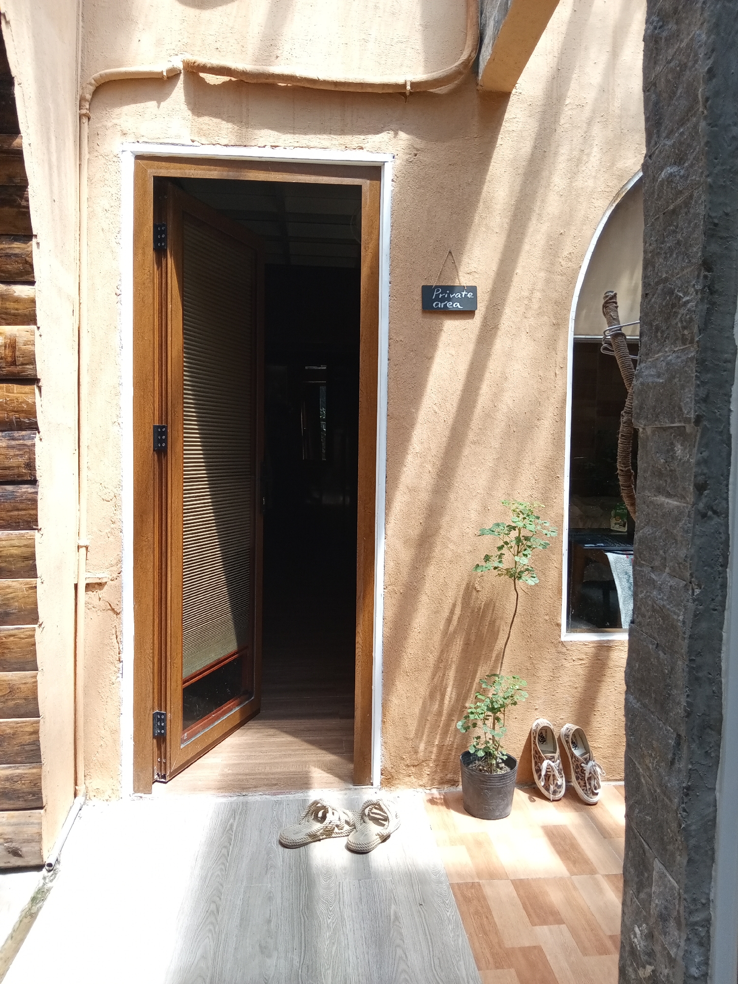 A photograph captures an exterior scene, depicting an entrance of a building with an open wooden door. To the left of the door, a wall constructed of logs is visible. Above the door, a beige stucco wall extends, with a small, framed sign reading "Private area" hanging to the right. Next to the sign, a window is framed in the same beige stucco, and a potted plant and two pairs of shoes are arranged below it. The floor is tiled in alternating light and dark brown shades. The scene appears to be located in the Bac Natural Heritage Area Trang, Vietnam, based on architectural and material styles.