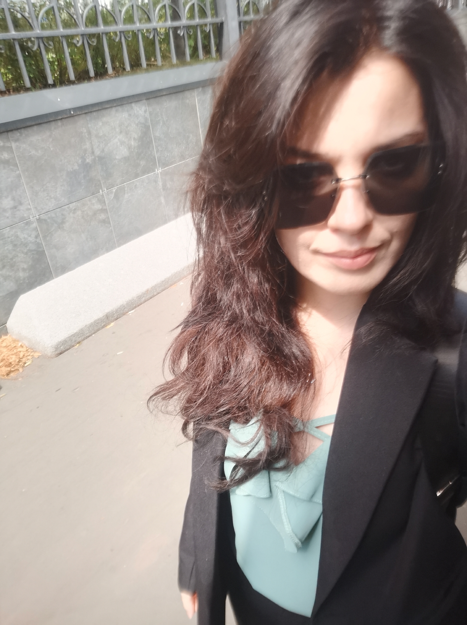 A woman wearing a black blazer and a light blue top with a criss-cross neckline is taking a selfie outdoors. She has long, dark brown hair and is wearing large, dark sunglasses. The background appears to be a city street with a grey tiled wall on the left, a metal fence with greenery behind it, and a paved area. There are some fallen leaves on the ground. The lighting suggests it is daytime. The environment is consistent with an outdoor urban setting, possibly in Moscow, Russia, given the architectural style of the fence and the prevalence of grey tiling in public spaces there.