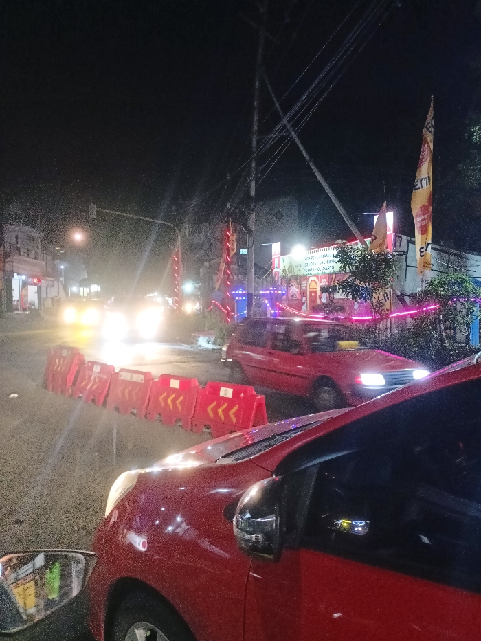 The image presents a vibrant nighttime street scene in Mojosari, Indonesia, captured from the vantage point of a red car, whose hood and side mirror are visible in the foreground, speckled possibly by rain or dust. The setting is an urban road or intersection, characterized by artificial lights against a dark sky.
In the mid-ground, a line of red plastic road barriers, marked with white and yellow chevrons ("<<<"), partially obstructs a lane, indicating traffic control or a designated area. Behind these barriers, a red sedan or hatchback approaches, its headlights cutting through the darkness. Further down the road, several other vehicles are visible as bright blurs of light from their headlights, suggesting ongoing traffic. A distant streetlight and a traffic light are also faintly visible.
Along the right side of the street, a building is brightly lit with festive pink and blue decorative lights. A prominent sign on this building reads "POS PENGADUAN KASUS KORUPSI" (Corruption Case Complaint Post), with additional text below including "KAWASAN ESTADION DALAM..." and "DORSEMOJO." Tall banners and flags adorn the area; a large yellow flag displaying "EMI" is visible on the far right, and several red banners with gold or yellow accents are also present. Overhead, a complex network of power lines crisscrosses the dark expanse of the night sky. The scene depicts a lively, illuminated street at night, possibly bustling with activity or celebrating an event, under typical urban conditions.